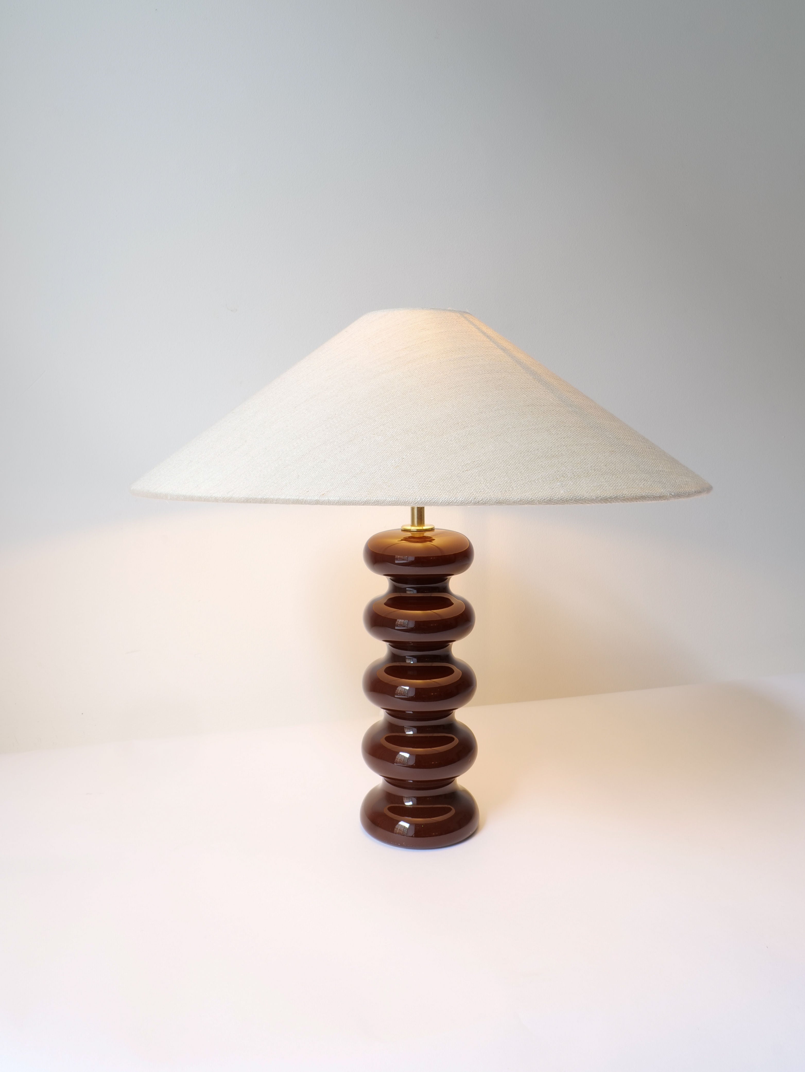 The Sculptural Ceramic Lamp by Collection apart showcases a stacked circular base in an elegant bitter chocolate color, crafted by hand in Sweden. It is paired with a wide, cream-colored fabric lampshade, all presented against a plain white background.