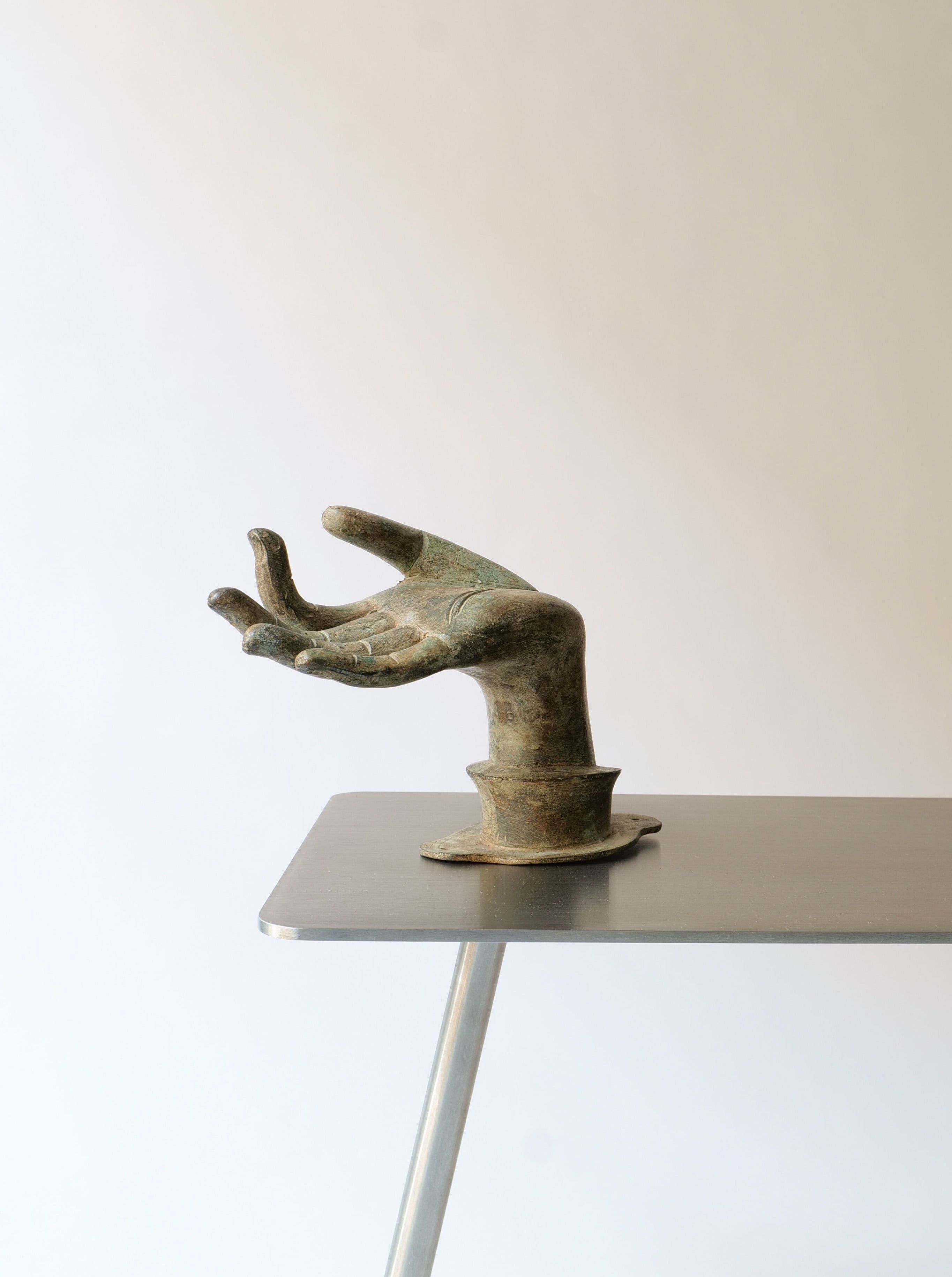A bronze sculpture of an open hand stands upright on a minimalist metal table. The plain background creates a stark contrast, which highlights the intricate details and emotional power of the elegant Bronze Hand by Collection apart.