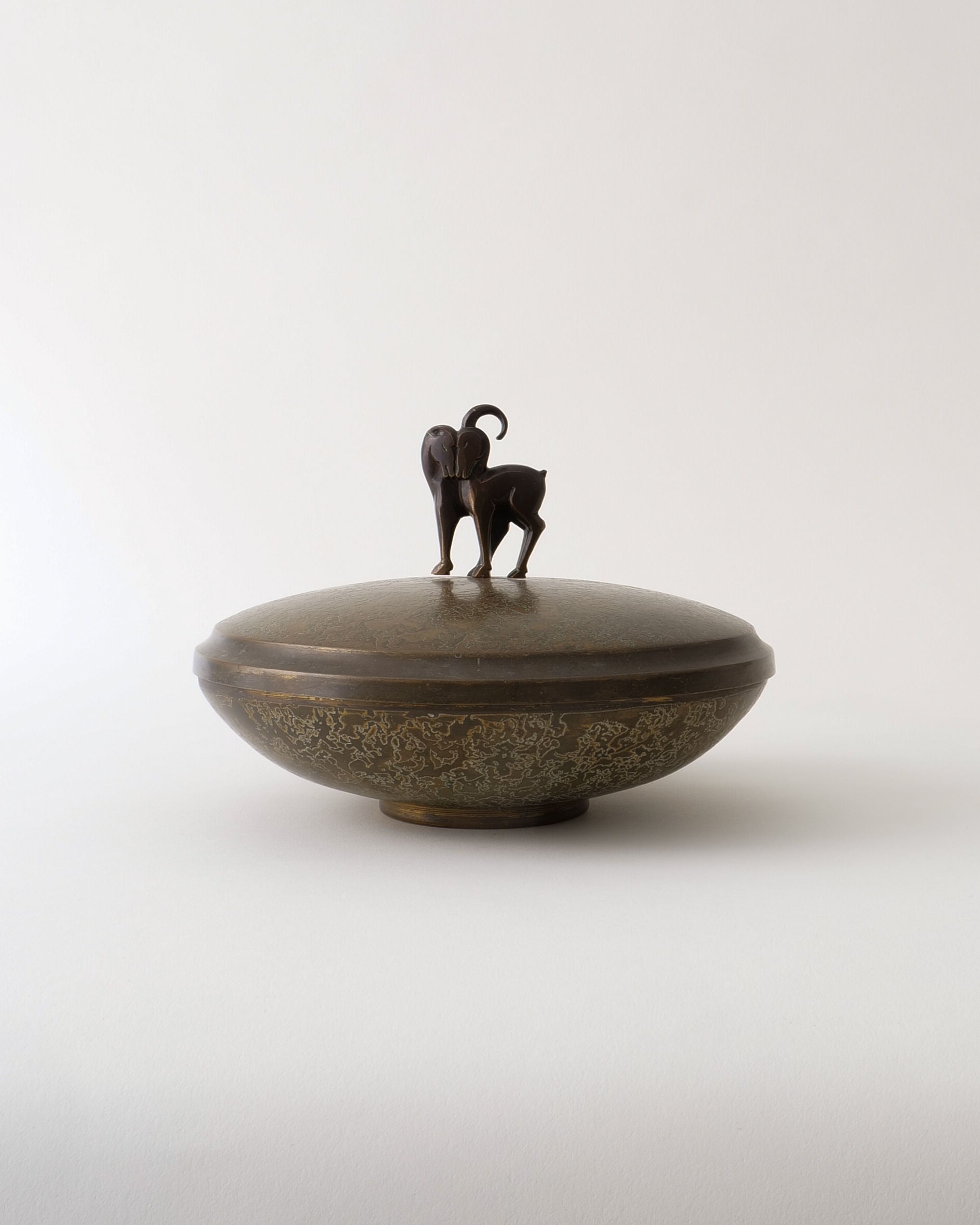 The Deco Box by Pierre Amédée Plasait for Collection apart is a round bronze lidded container featuring delicate patterns and a sculpted goat atop the lid, showcasing early deco craftsmanship against a plain light background.