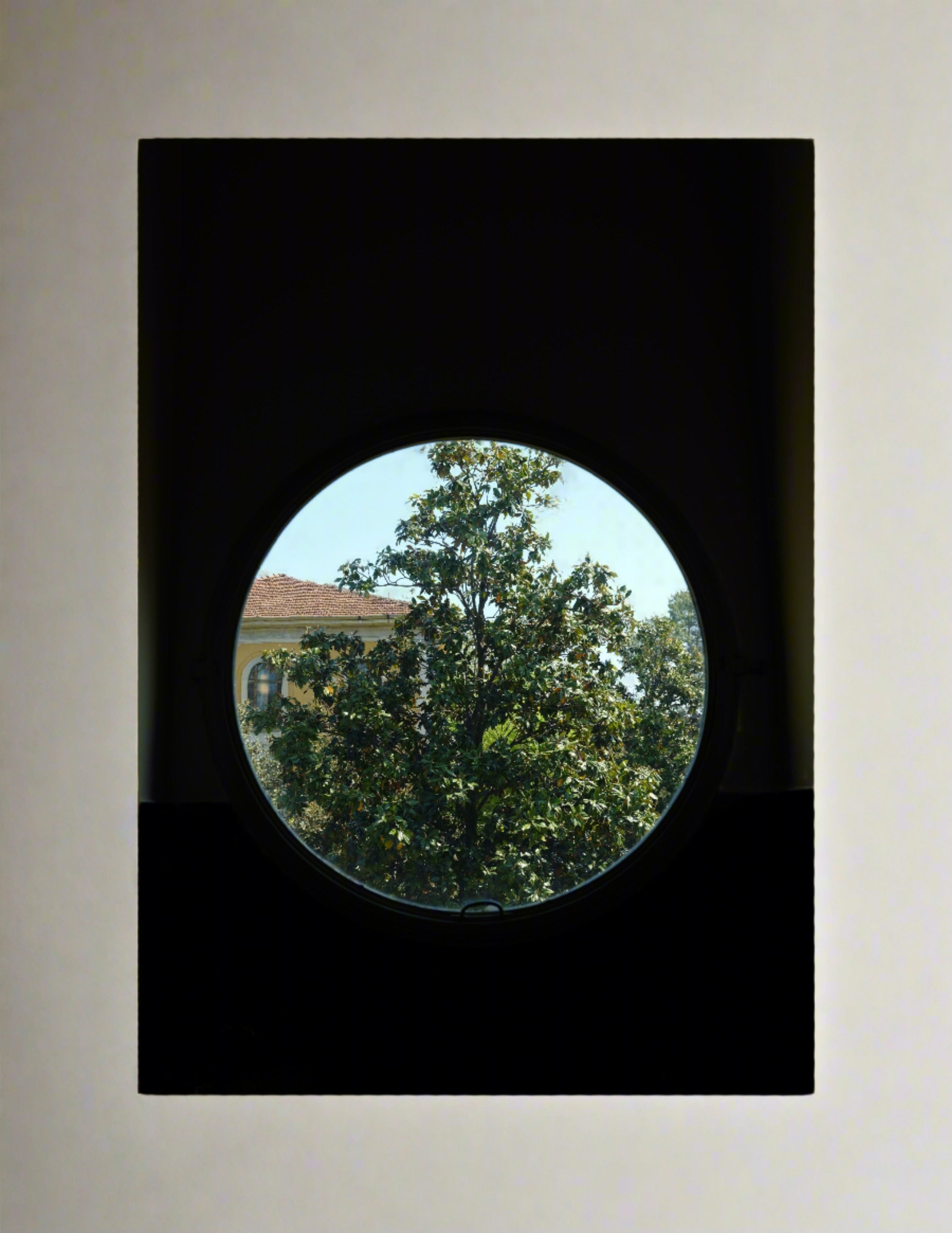 A circular window set in a light-colored wall frames a picturesque view of a leafy tree and a distant building with a reddish roof. This Grace Rose Watts photography captures the composition, contrasting the dark interior with the bright, natural scene outside. Available as "Villa Necchi Campiglio, Milan - Print Photography" limited edition print.