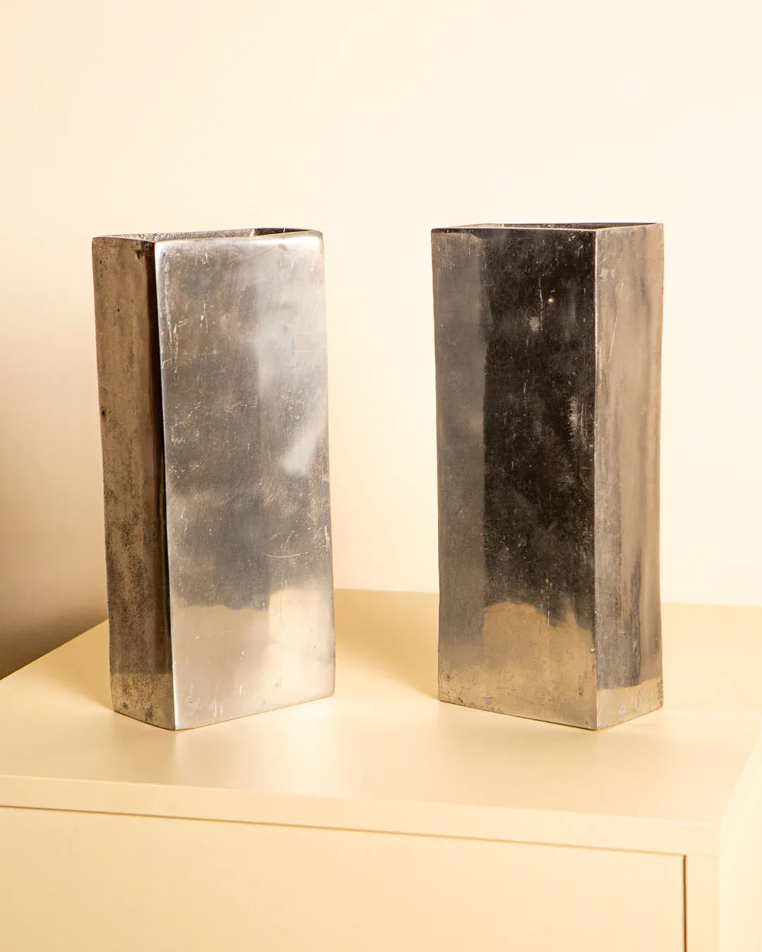 The Pair of 80's Italian Aluminum Vases by Treaptyque, tall and rectangular with a reflective surface, are elegantly placed on a light tabletop against a neutral wall, highlighting their minimalist and modern Italian craftsmanship.