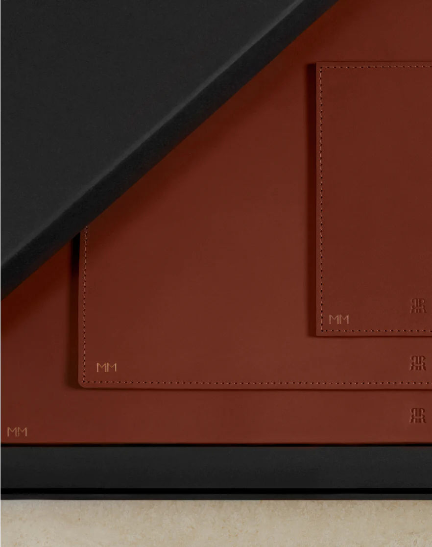 Corner view of a Paradise Row Leather Home and Away Gift Set's desk mat, showcasing black and brown vegetable-tanned leather with stitched edges. The brown part has "MM1" and "RR" engravings at the bottom right, while "MM1" appears on the black section's bottom left.