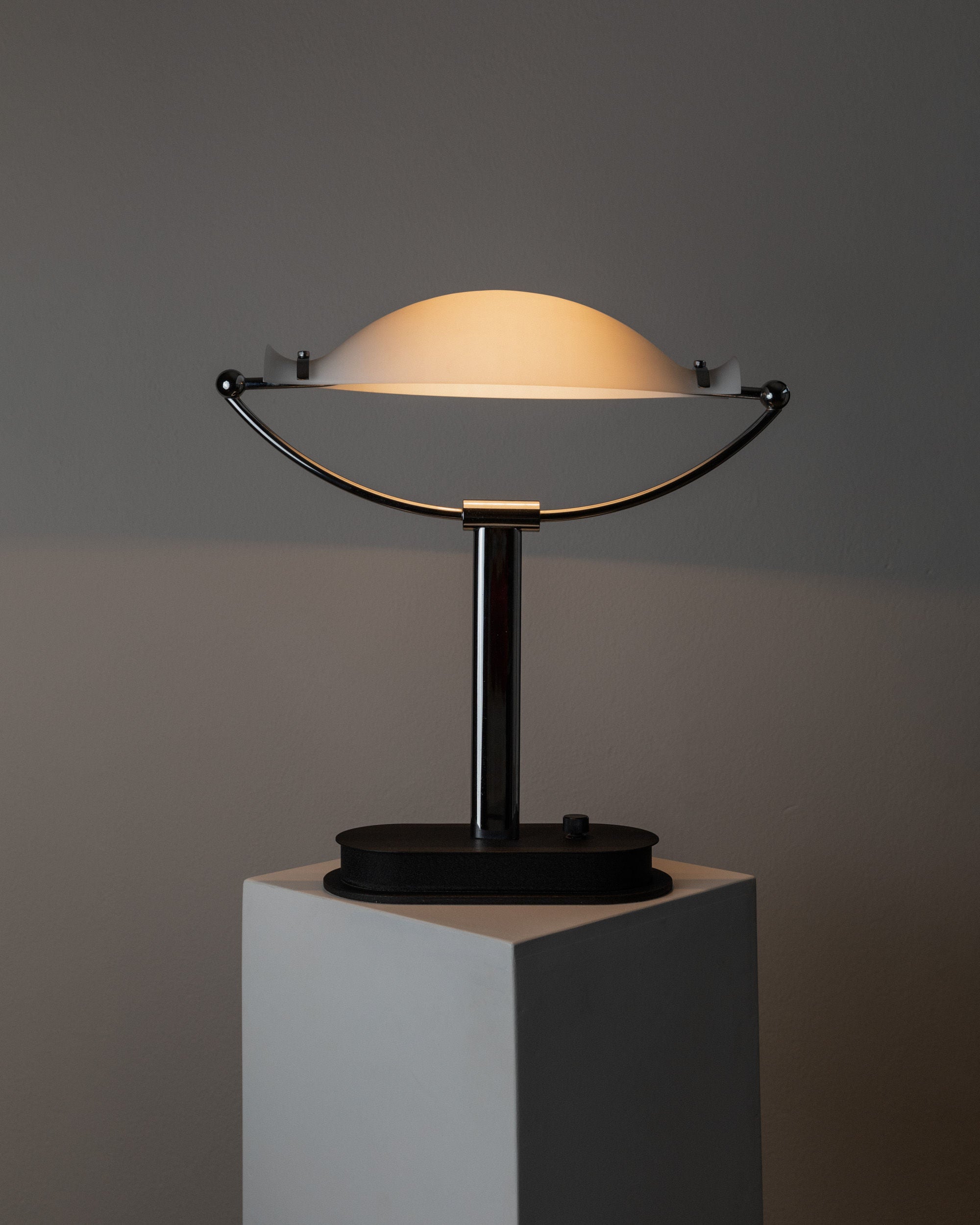 The Murano Glass Table Lamp by Bottega Jacobs features a semi-circular glowing shade on a rectangular pedestal. Its sleek metal design exudes retro charm, casting soft light against neutral walls, reminiscent of 1970s space age style.