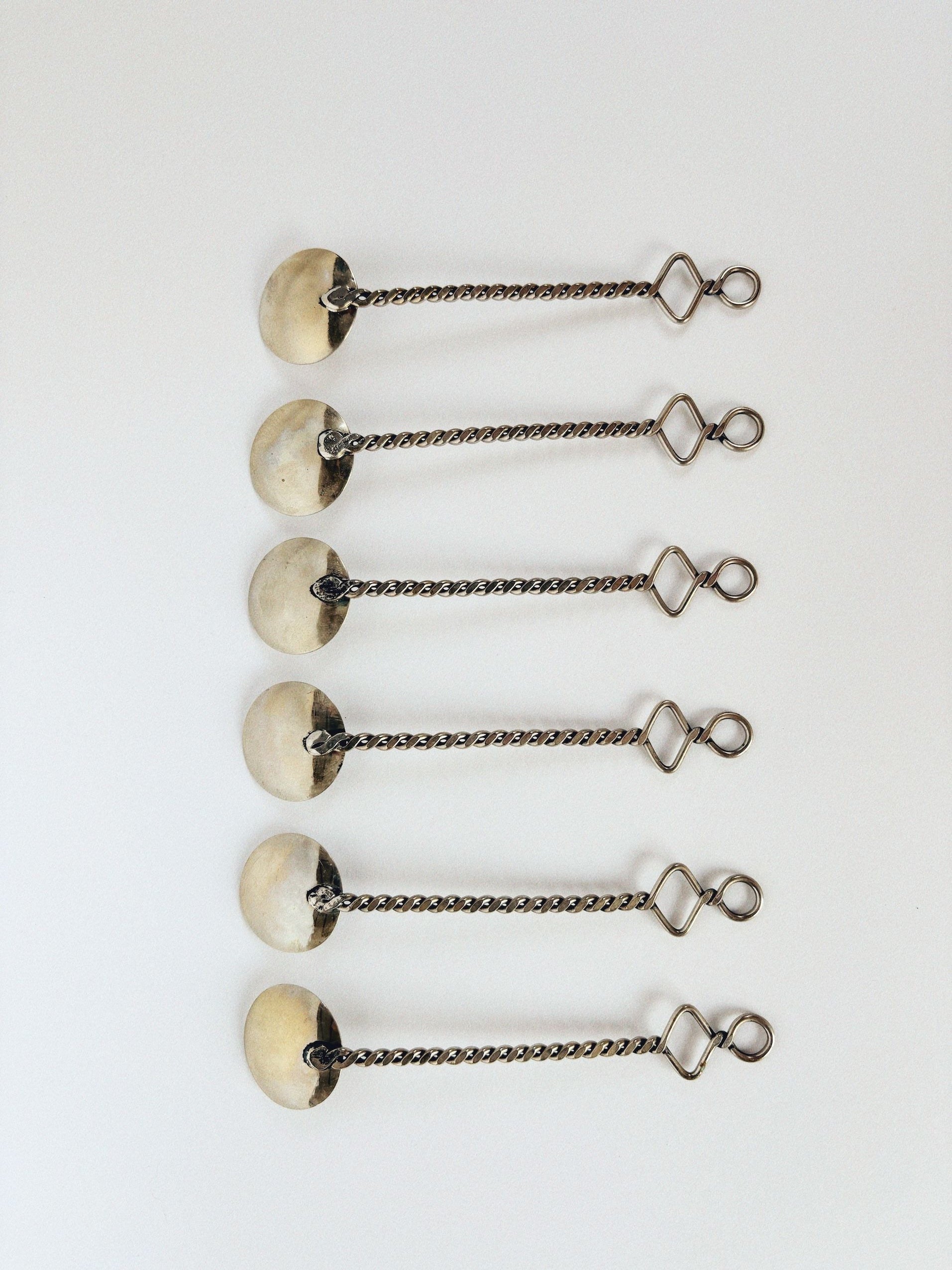 Six identical items, like functional art objects, are aligned vertically on a white surface. Each features a circular disk and a lobster clasp linked by a twisted chain segment, similar to the 70's Handmade Spoons Set by Huerto.