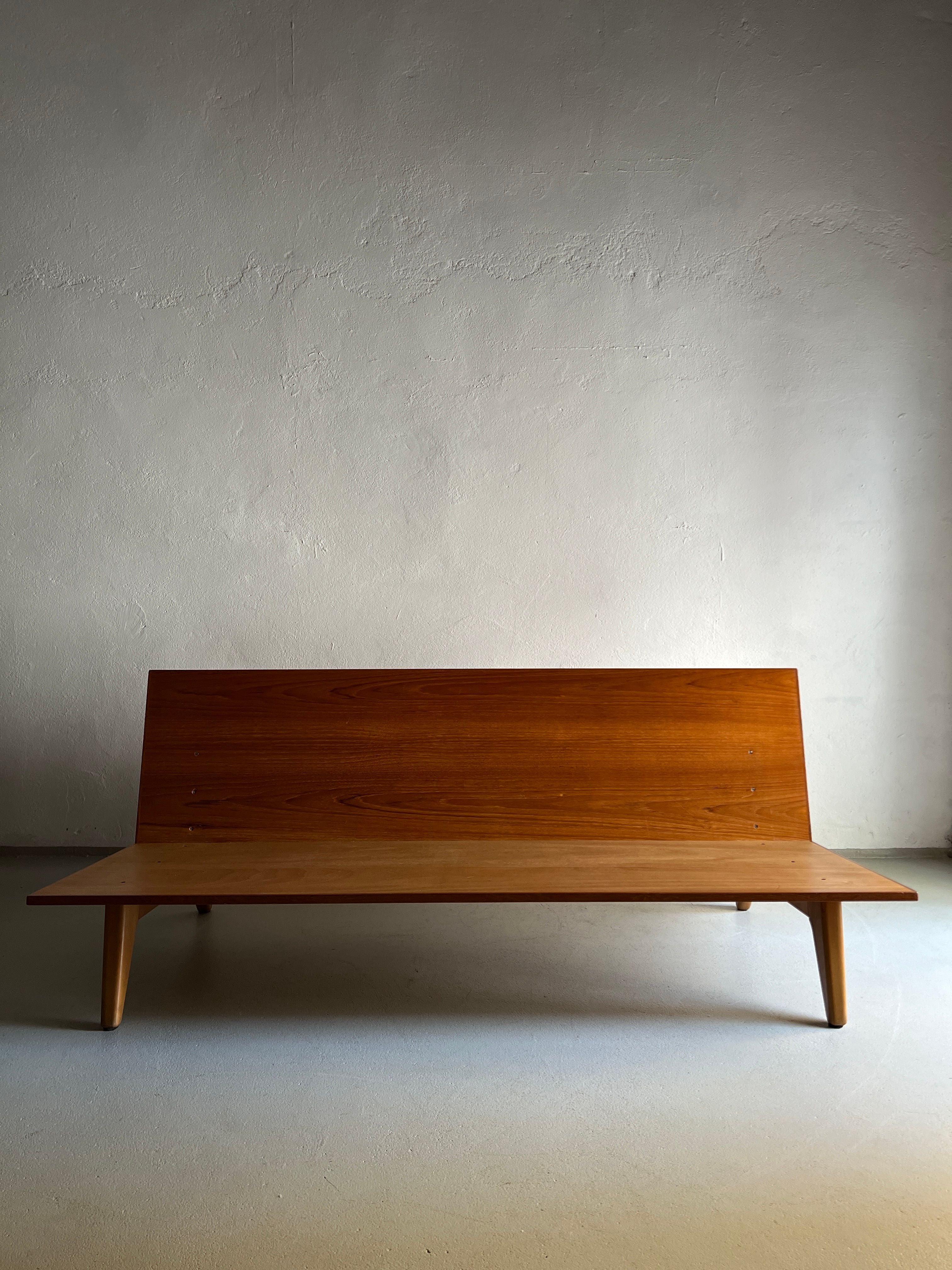 A minimalist wooden bench crafted from teak boards stands against a light-colored, slightly textured wall. The **Teak Daybed Sofa by Gustaf Hiort Af Ornäs | Finland | 1950s** by **Veter Vintage** has a slanted backrest and tapered legs, creating an elegant and simple aesthetic. The setting appears to be indoors with soft, natural lighting.
