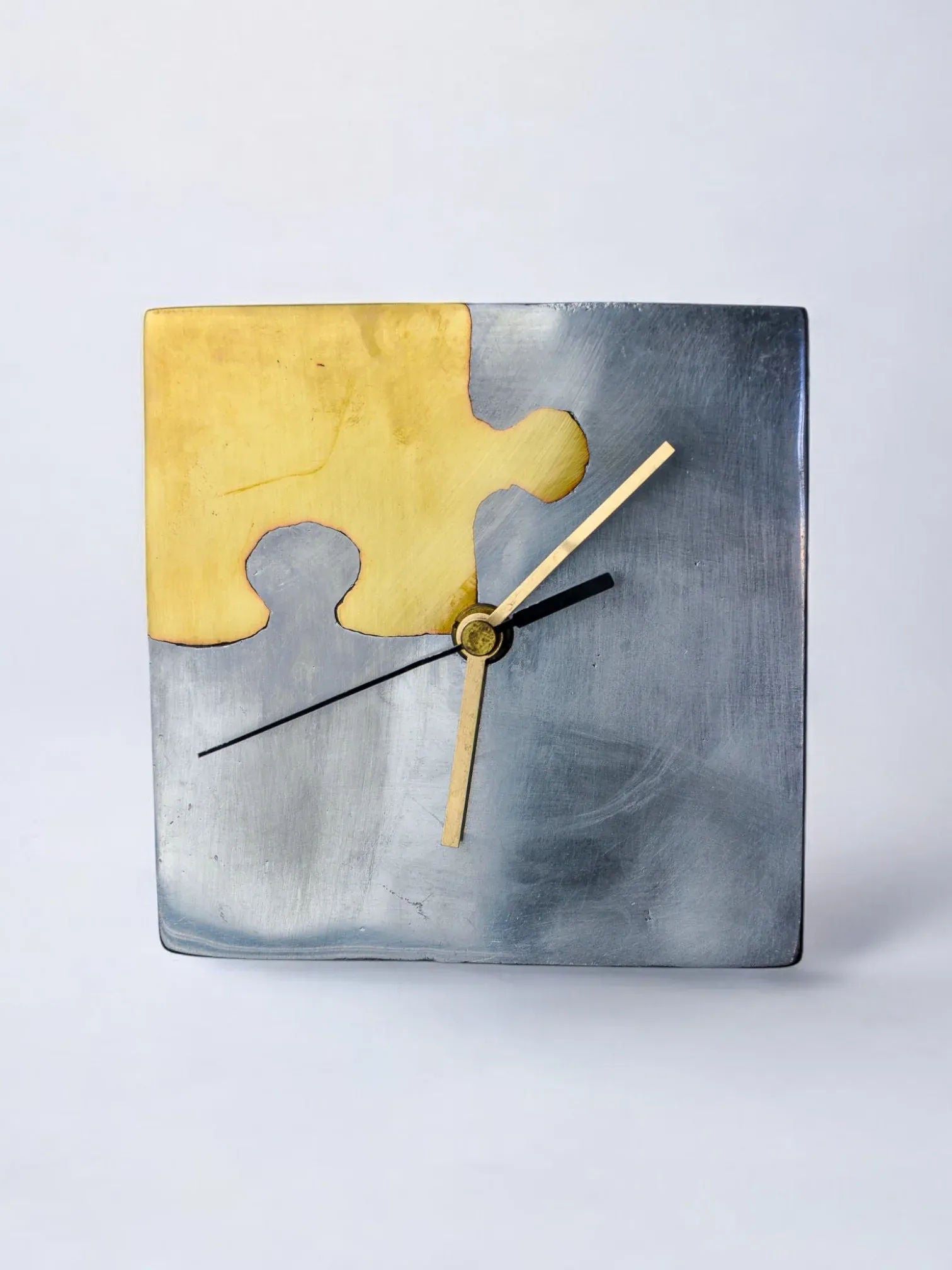 The Brutalist Clock "Puzzle" by Malamar Studio, Spain, 1980, features a square design with metallic silver and gold overlapping puzzle pieces. Its minimalist black and gold hands on a light background make it an eye-catching piece for any modern space.