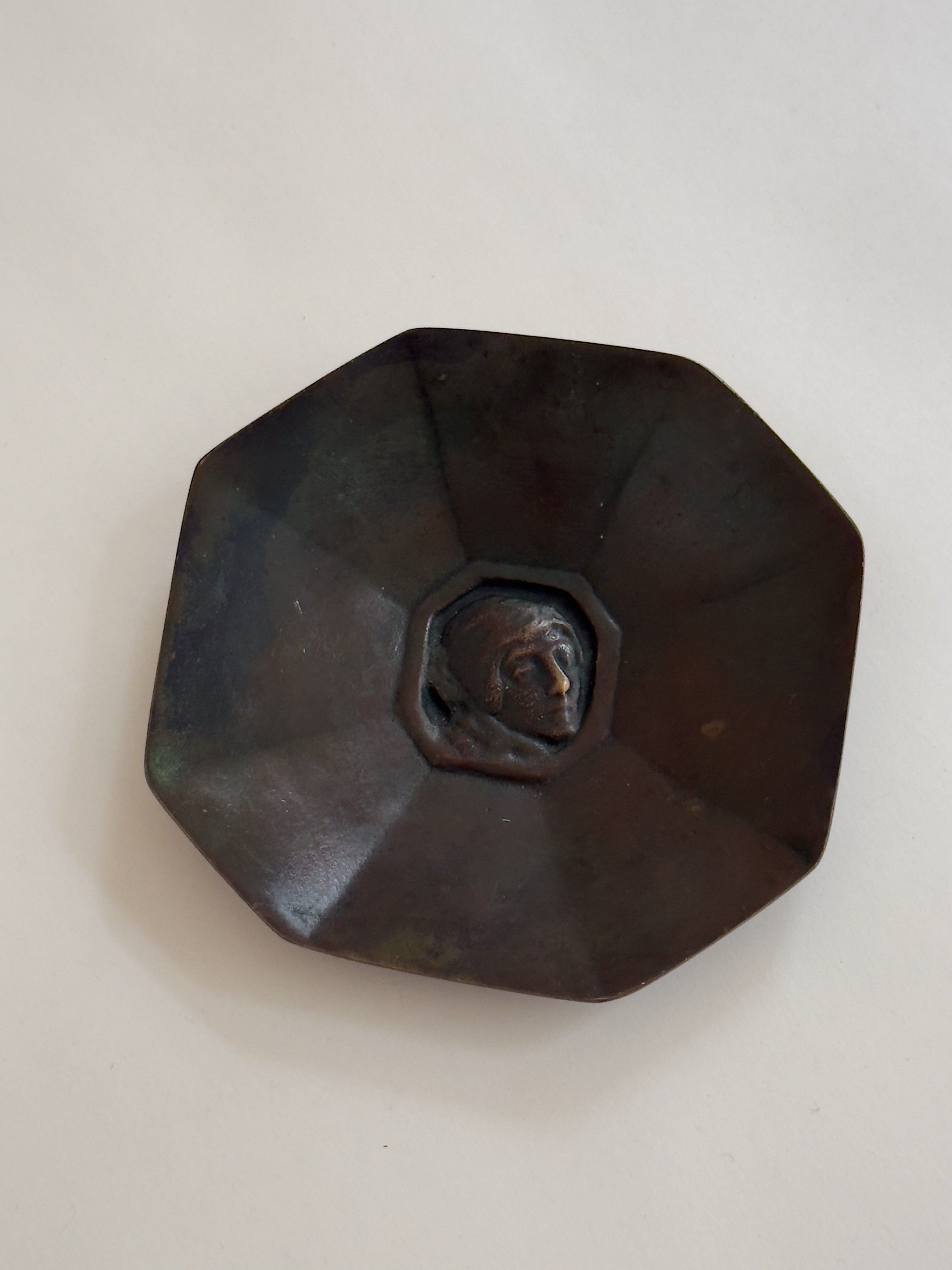 The Octagonal Bronze Dish by Jacob Ängman, GAB (Galerie Storm) is a small, dark bronze piece featuring a raised head in profile at the center, viewed from above on a plain light background.