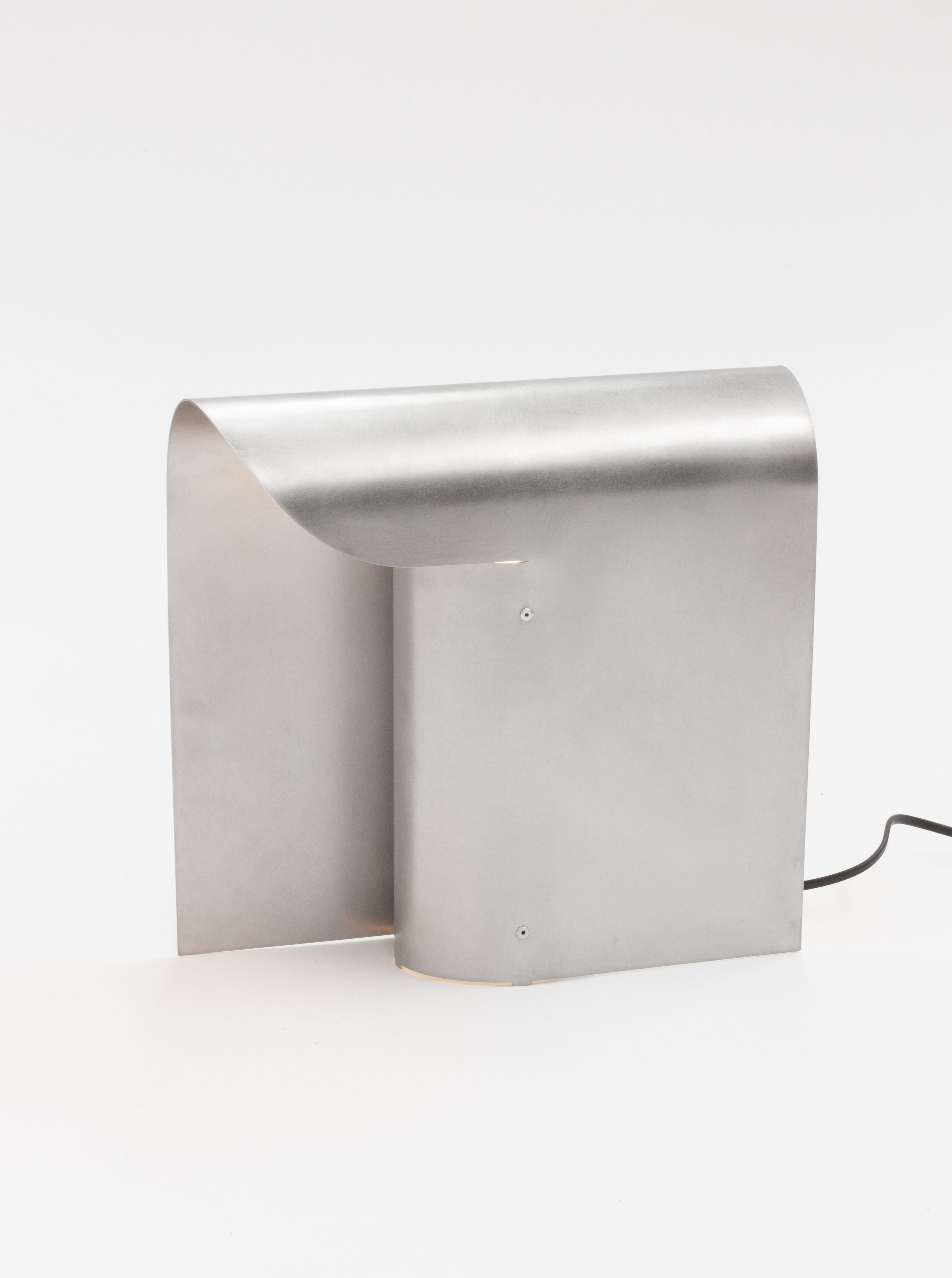 A sleek, modern metallic light fixture with a minimalist design, featuring a curved sheet of brushed stainless steel standing upright. The elegant and contemporary appearance is complemented by an E27 socket for versatile bulb options, with a black electrical cord partially visible on the right side. This is the Arc Table Lamp by Paul Coenen.