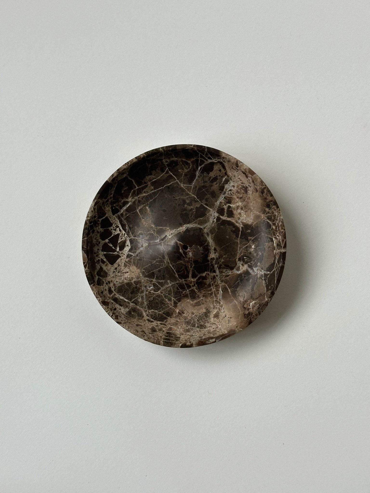 The 'ALTAR' Incense Holder by CARRARA Studio is a round, shallow dish handcrafted from dark brown marble with light tan and white veining—an elegant solid stone accent for your space.