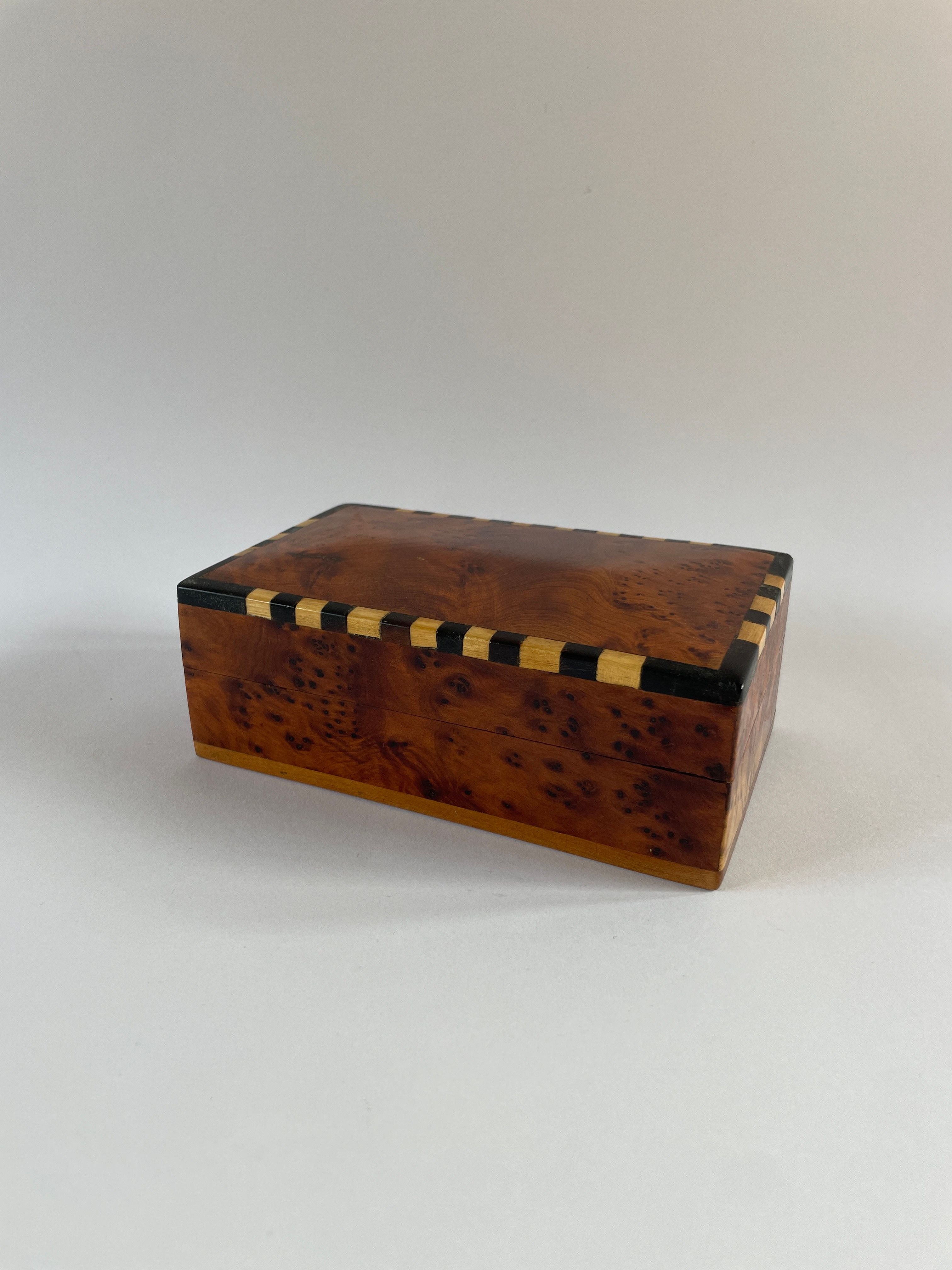 Introducing the Art Deco Burr Box by Maud Vaughan: a compact, handcrafted lidded box boasting a sleek polished finish. Its lid is adorned with an art deco-inspired checkerboard pattern along the edges, providing a striking contrast to the smooth, natural brown wood grain. The surface is accented with unique black speckles for added character.