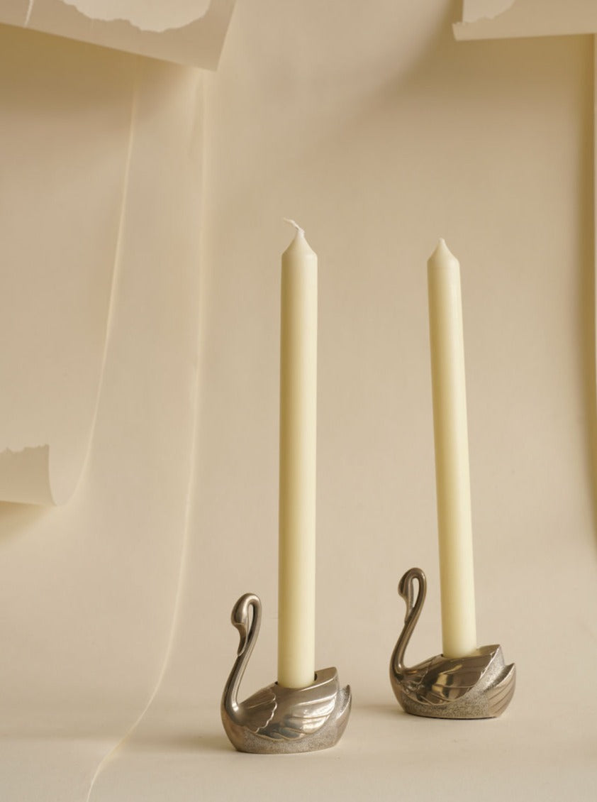 A pair of white taper candles are placed in delicate Les Objets Amsterdam Two Swan Candleholders against a beige background with draped fabric. The candleholders have a sleek, minimalist design, adding a touch of sophistication to the setting.