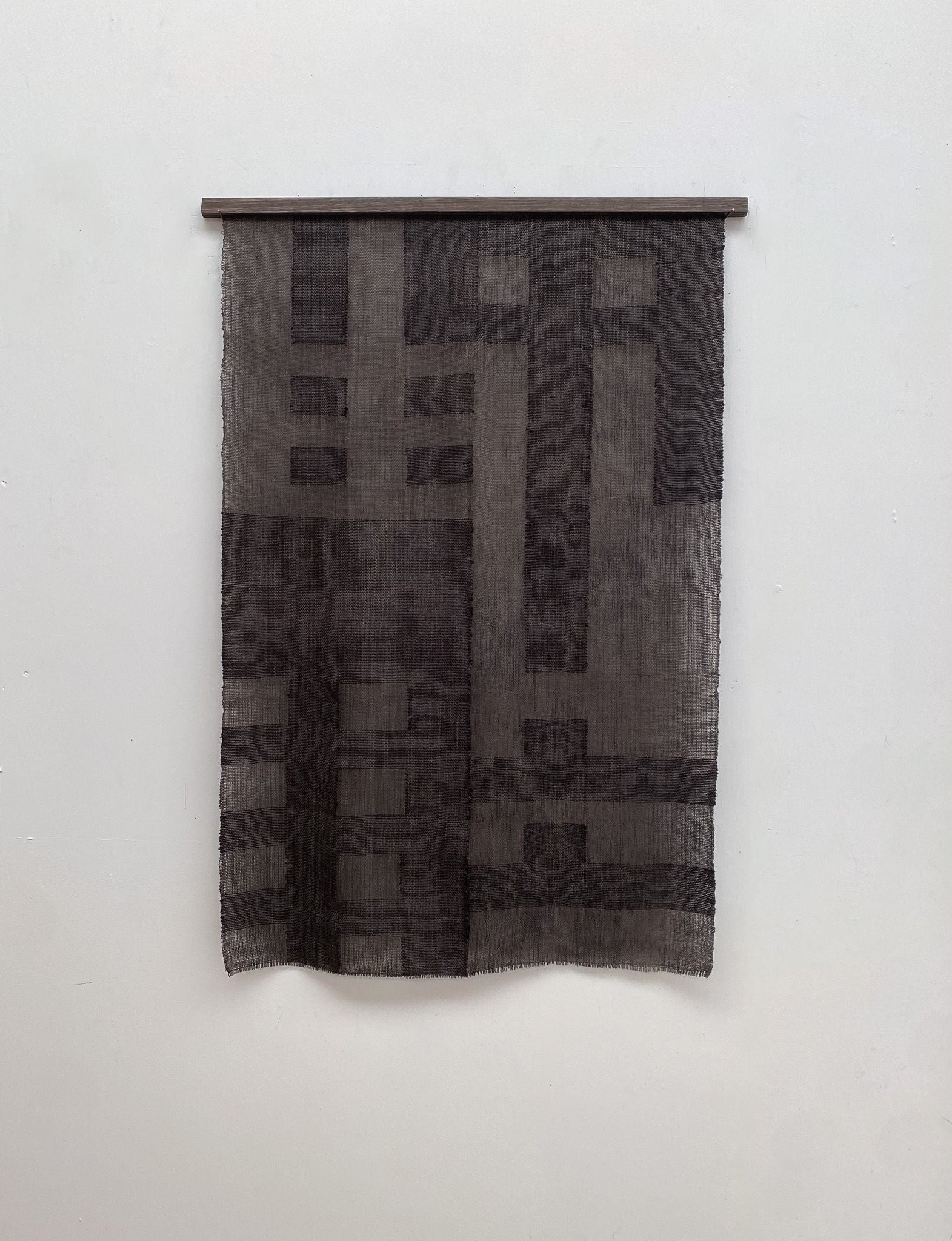 The Grid I tapestry by Sundance Studio, inspired by the Bauhaus movement, hangs on a wall. It features geometric rectangles and lines in textured gray for a modern, minimalist look.