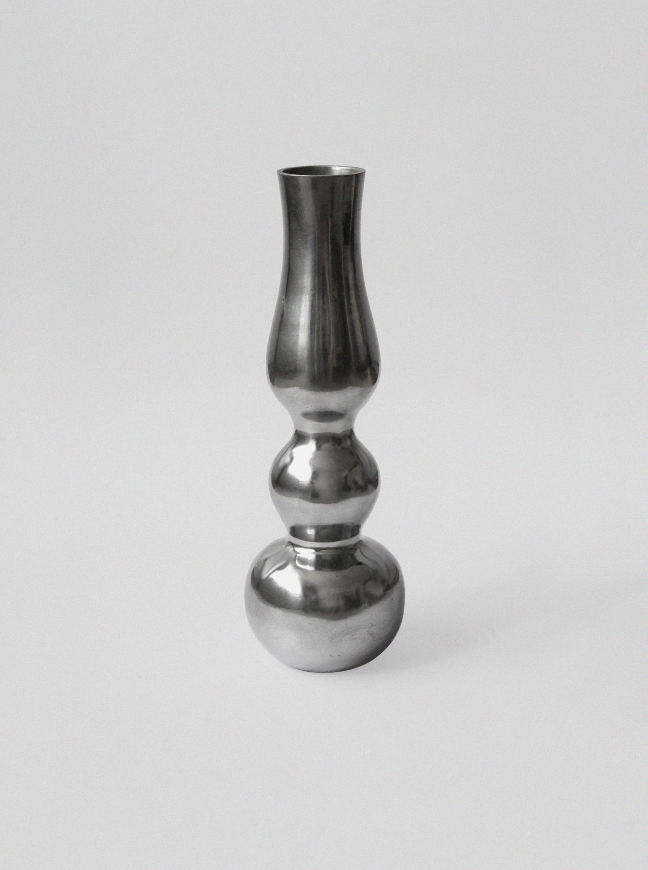 The Aluminum Figure Vase by Vintage Stories features a reflective, sculptural design with a mirror finish. It stands out on a plain light gray background, capturing the essence of minimalist style.