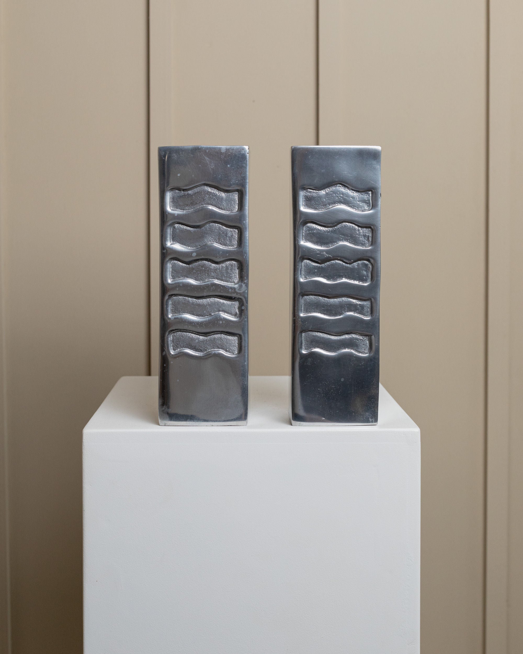 A pair of artistic metallic rectangular vases from the Bottega Jacobs Metal Vases Set Italy 1960s are displayed on a white pedestal against a beige paneled wall, featuring shiny, reflective surfaces with wavy patterns.