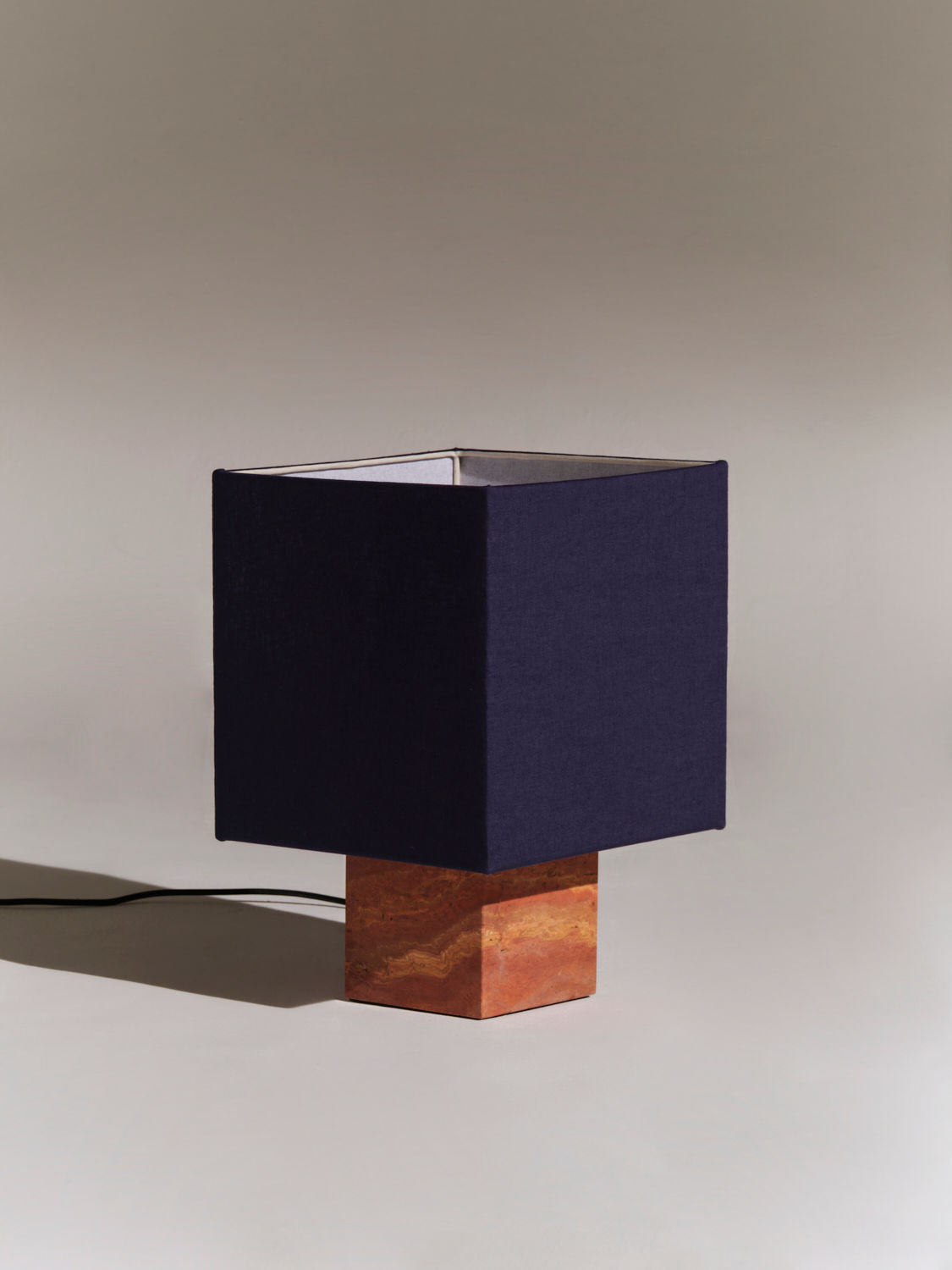 A modern table lamp with a square, navy-blue fabric shade and a wooden base made from natural materials is positioned on a neutral background. A black electrical cord extends from the lamp's base, emphasizing its minimalist and elegant design. The Roma I Squared Red Travertine by Nassi Lamps stands out as an exquisite piece in any setting.