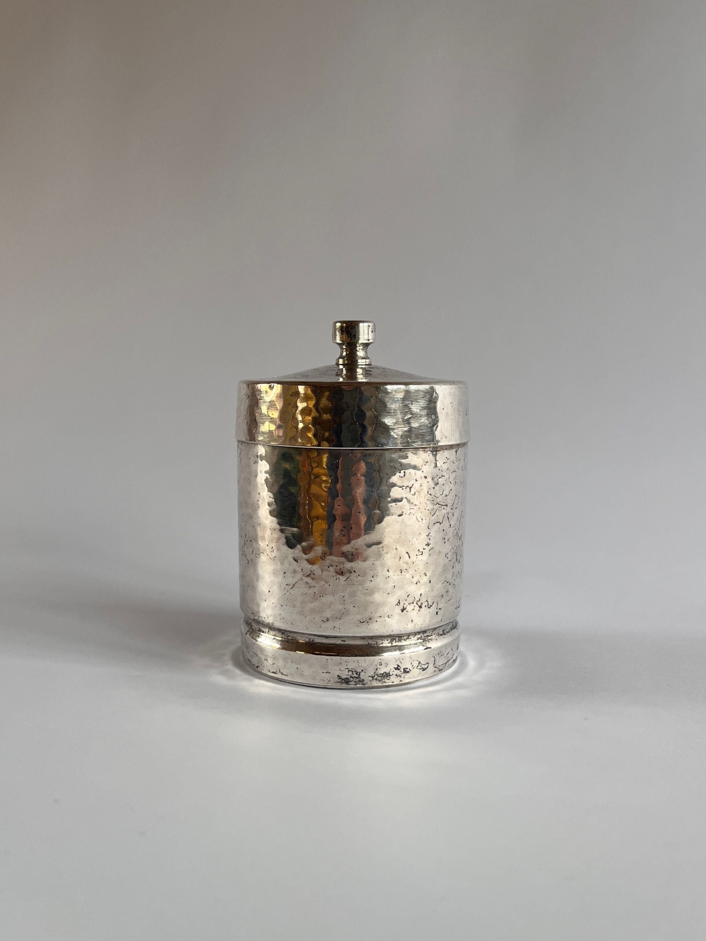 A small, cylindrical Art Deco Hammered Pewter Box by Gero from Maud Vaughan, featuring a lid with a knob, sits against a plain light-colored background, exuding elegance.