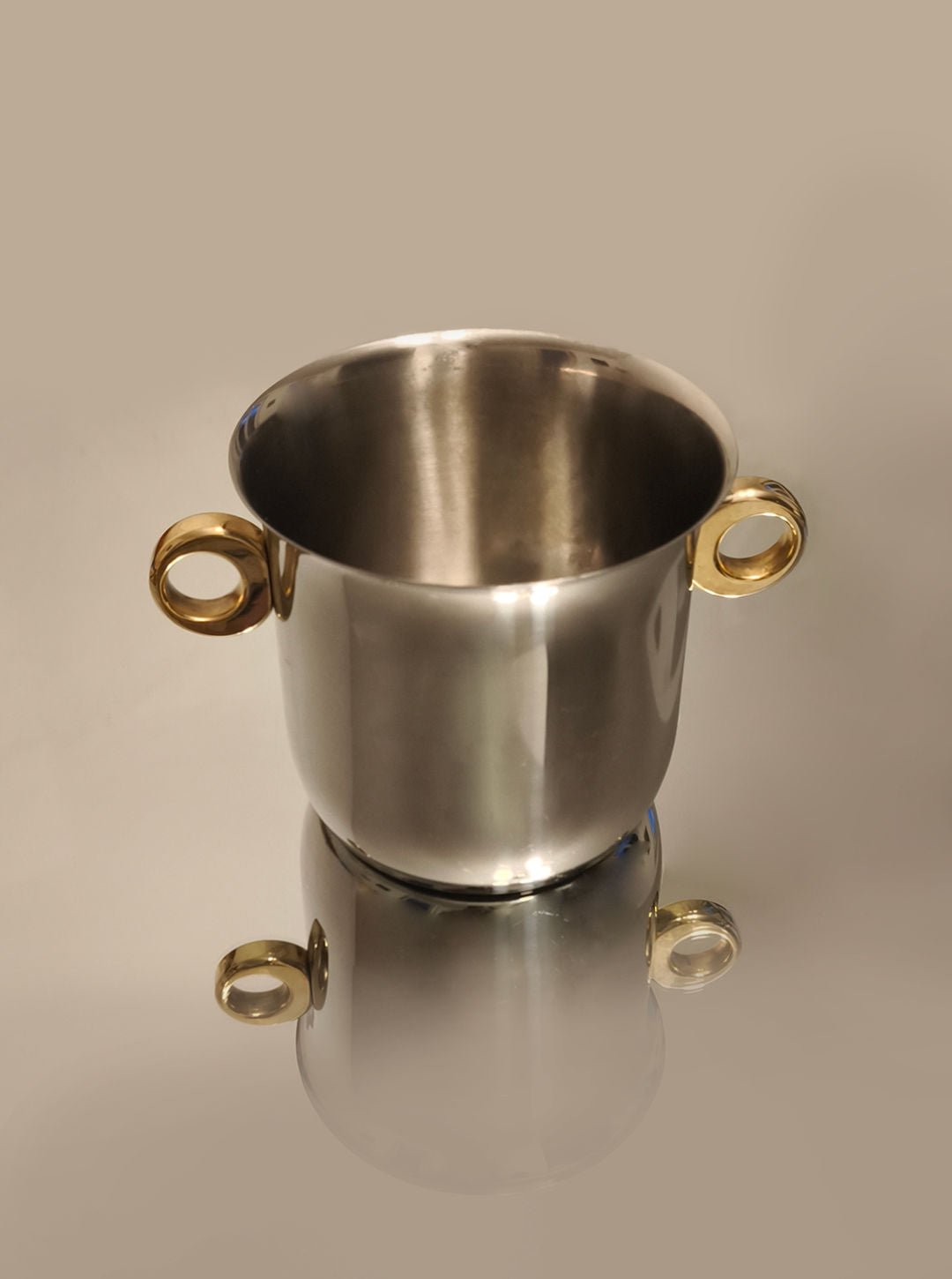 The Ice Bucket, Italy 1970s by Les Objoies, features silver stainless steel and two round brass ring handles. Its Italian modernist design sits on a reflective surface, casting a mirrored image against a light beige background.
