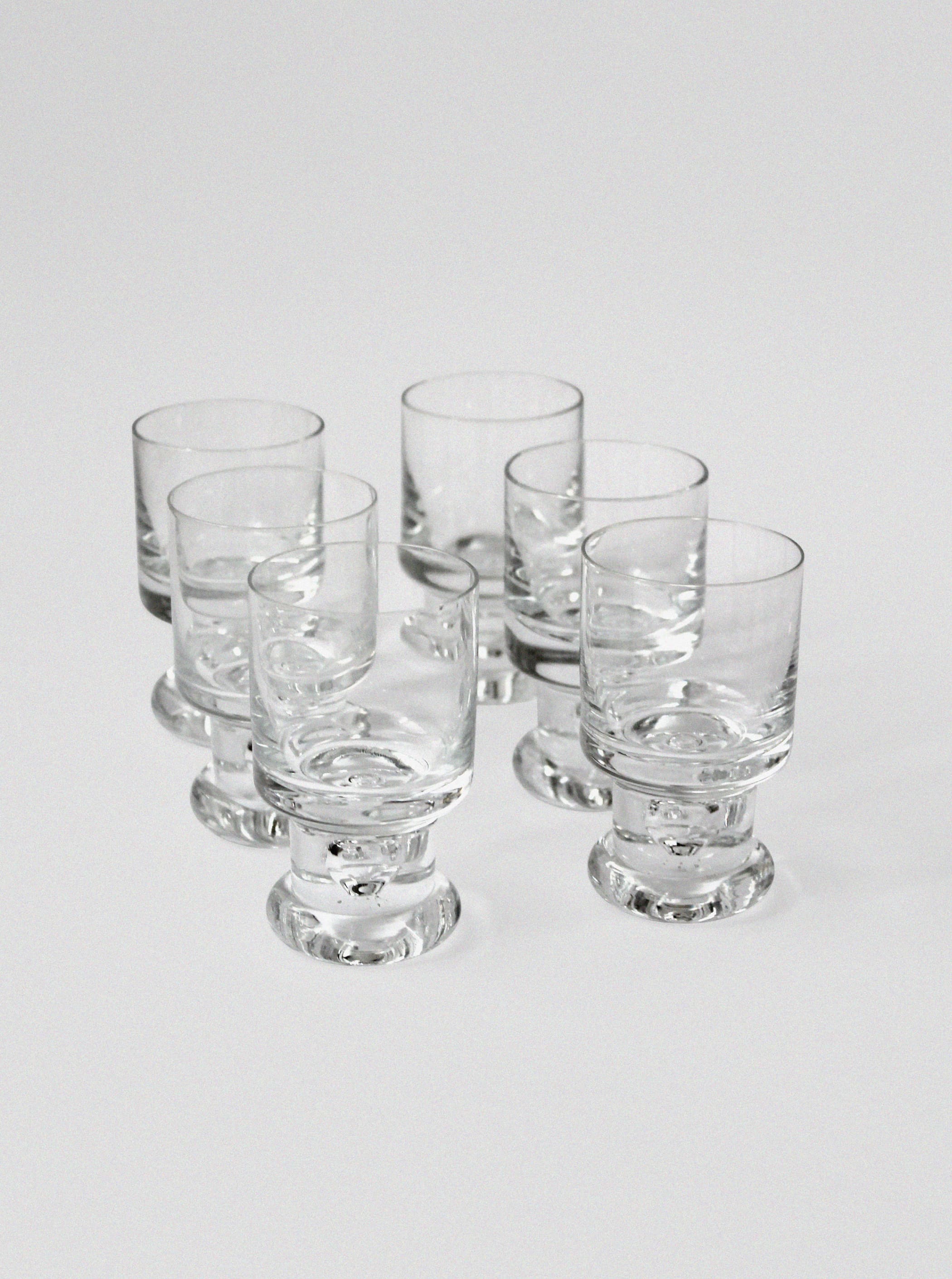 The Tapio Wirkkala Set of Glasses by Vintage Stories features six clear, empty glass tumblers with thick bases in a loose cluster on a plain white background, evoking classic Mid-century style.