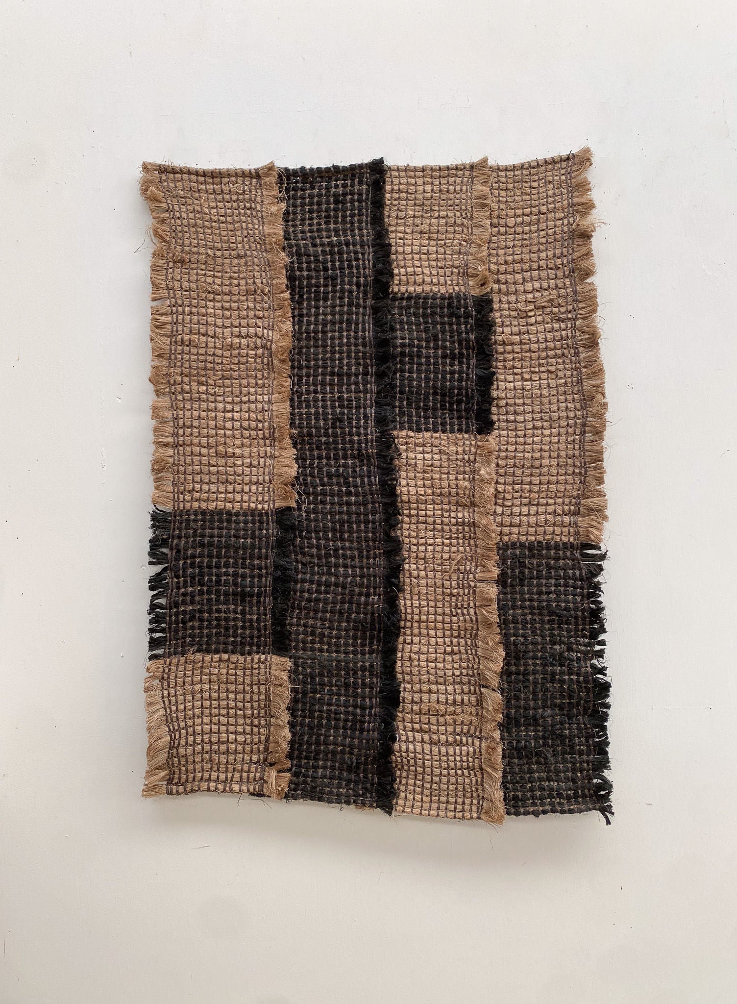 The Ink / Sand from Sundance Studio is a woven textile crafted from banana fiber, featuring a grid pattern with alternating light and dark brown sections. Its frayed edges enhance its rustic texture and highlight its biodegradable qualities.