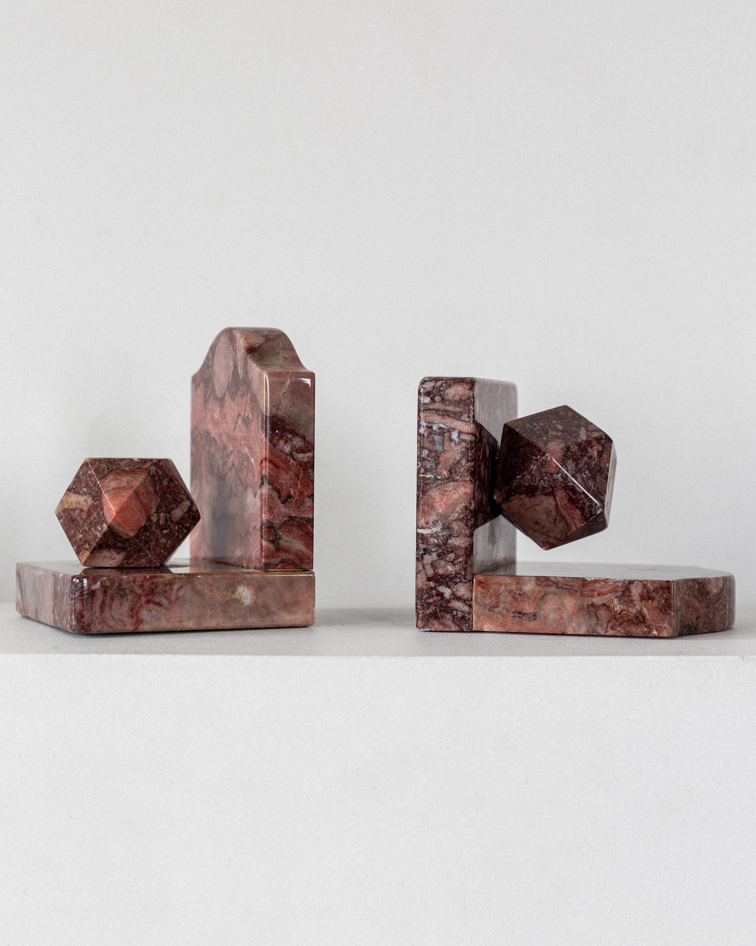 The Pair of Art Deco Marble Bookends by Spigel features two pieces made from marbled brown and red stone. Each bookend has a rectangular base with a unique 3D shape, reminiscent of abstract geometric sculptures, displayed on a plain white surface.