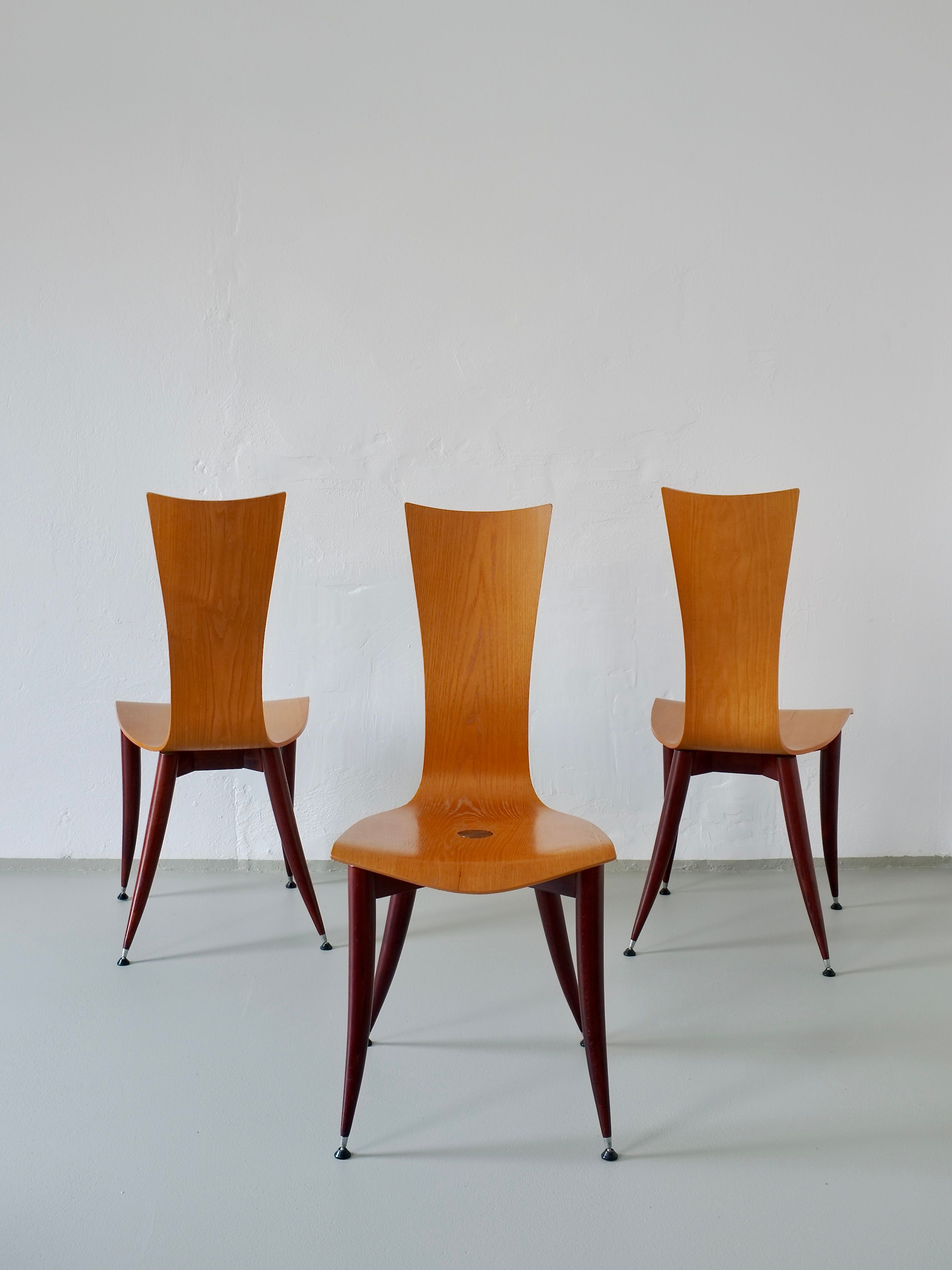 Three Veter Vintage chairs, with sleek curved backs and angled legs, are set in a white-walled room with a gray floor. These 1980s Italian plywood chairs have burgundy legs and a warm finish, exuding minimalist elegance reminiscent of Philippe Starck's style.