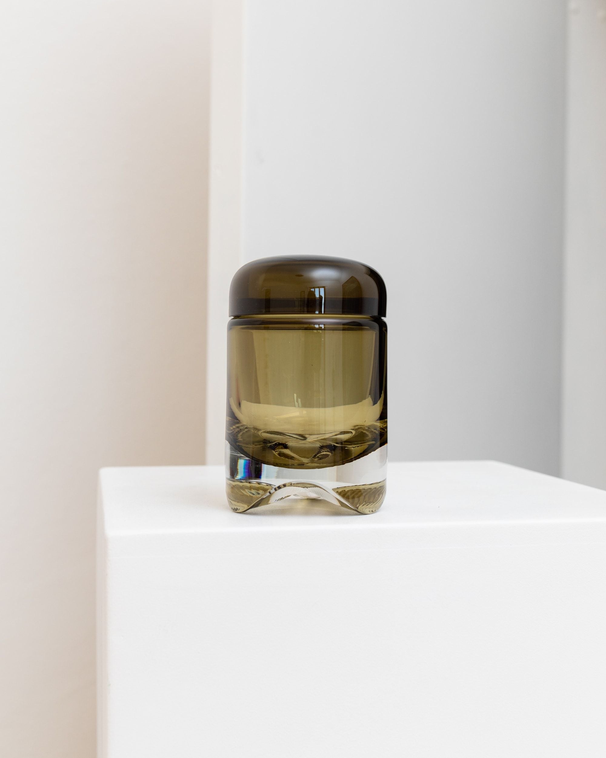A Vintage Glass Jar 1970s by Bottega Jacobs, encased in a small, dark glass bottle that mirrors a retro aesthetic, is perched on a white pedestal. Its minimalistic design and unobtrusive background emphasize the product's sophisticated look, evoking the charm of mid-century design.