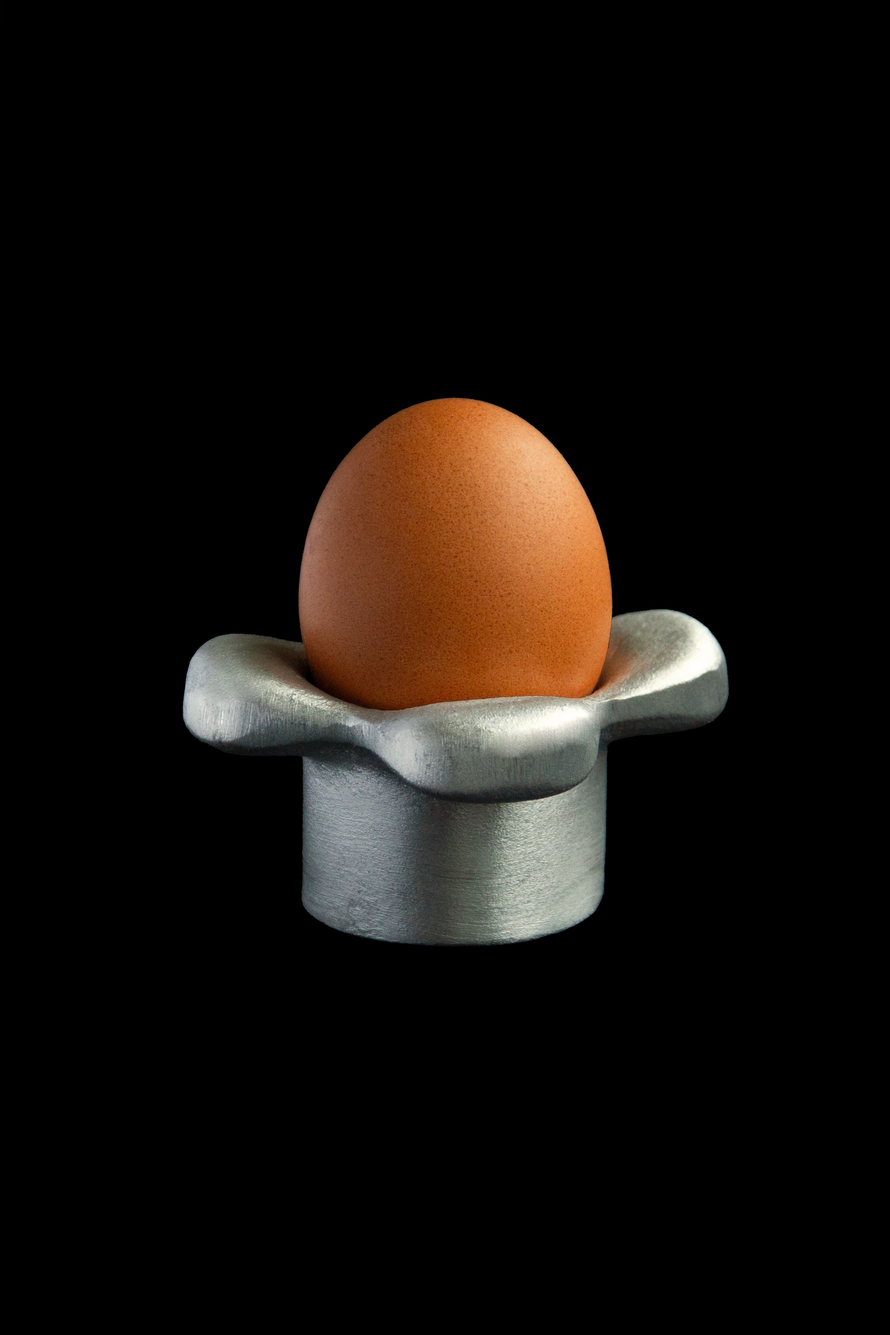 A brown egg stands in Atelier Barb’s shiny, upcycled aluminum 'Petal' Egg Holder, adding a sculptural touch against a solid black background.