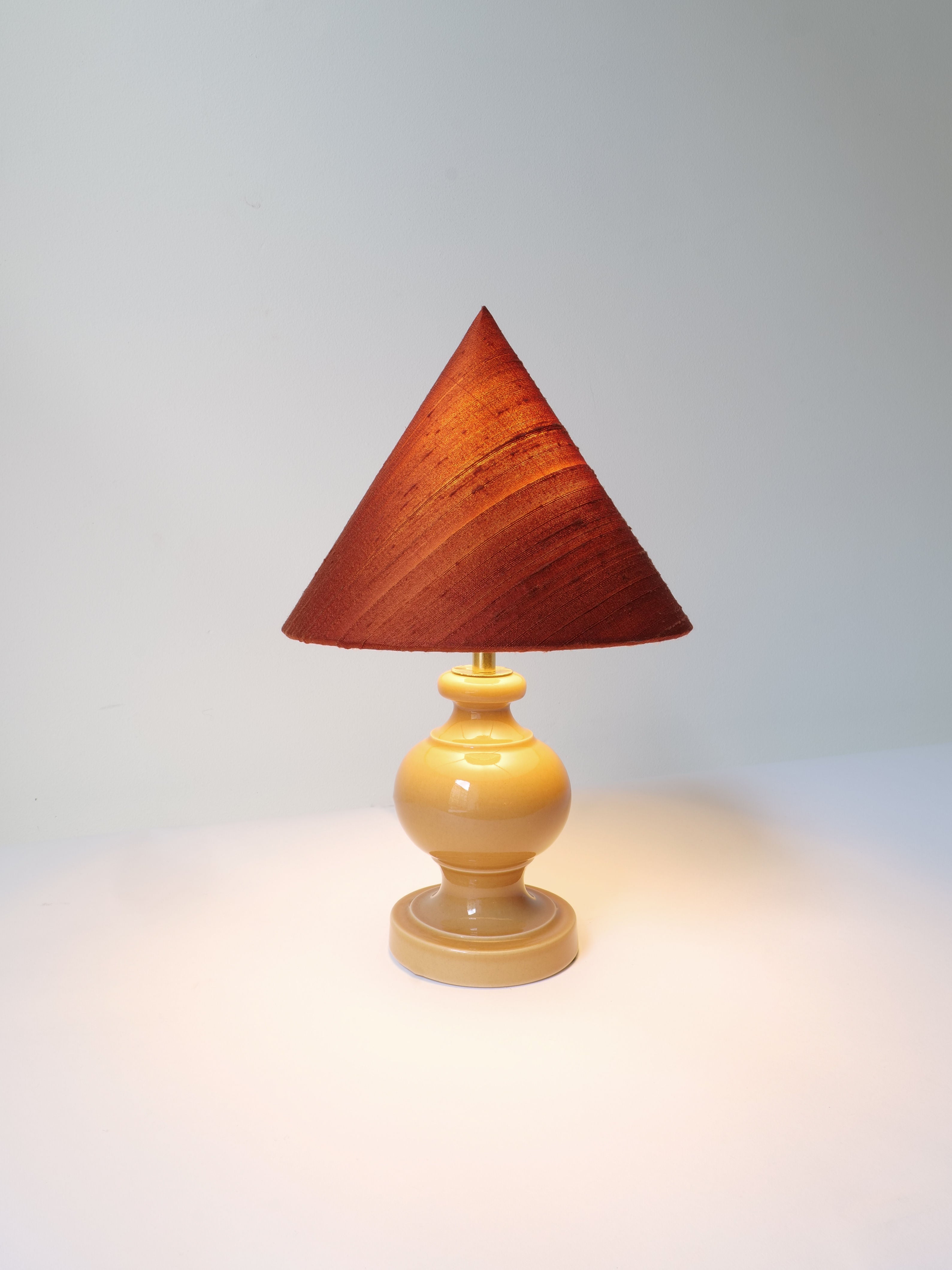 Introducing the Small Sculptural Ceramic Lamp by Collection apart. This lamp is a testament to simple elegance, featuring a beige ceramic base that echoes classic design sensibilities, paired with a wide conical brown shade. It is powered by an E27 lightbulb, which casts a warm glow across any white surface and wall behind it, creating a cozy ambiance.