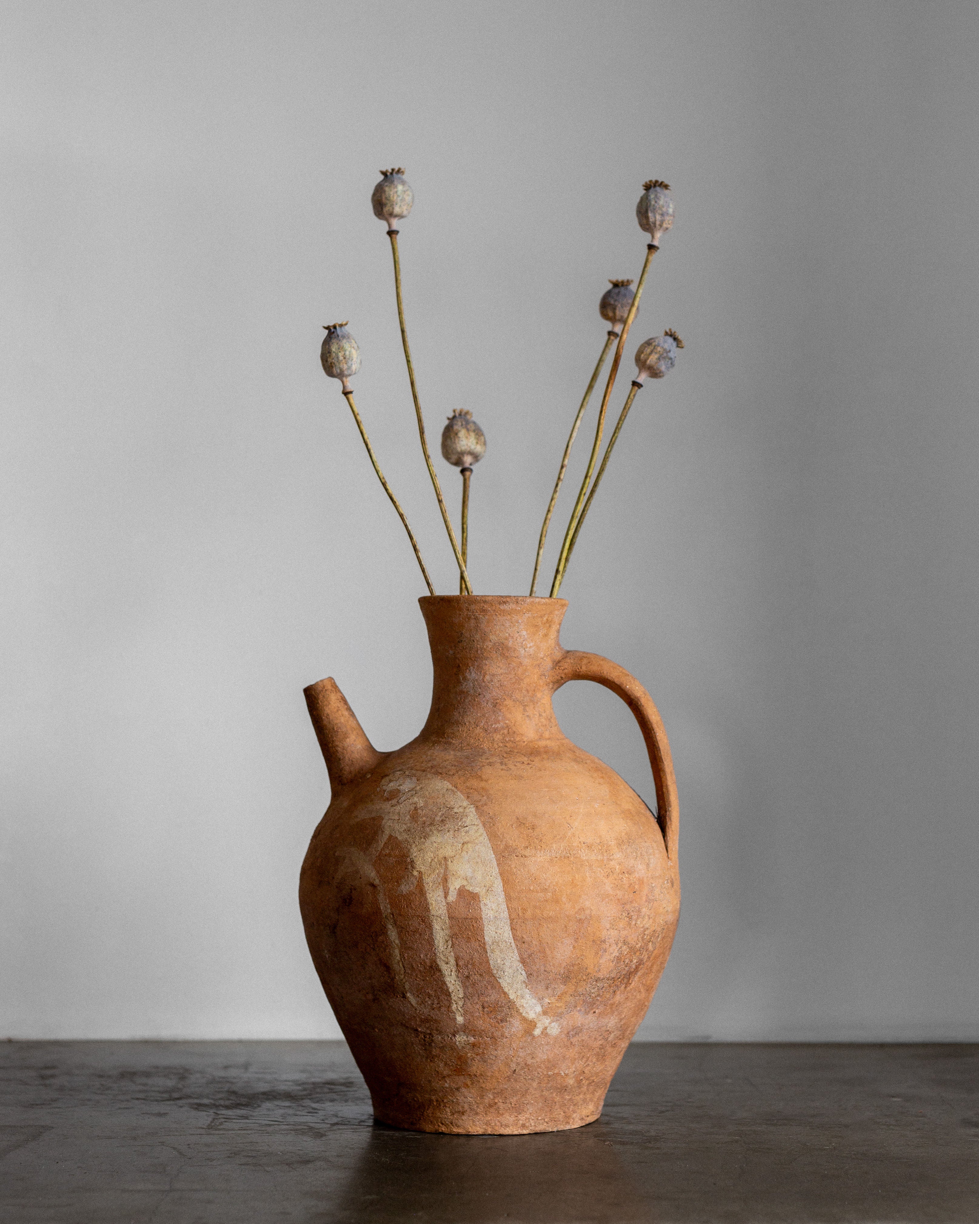 The Spigel Early 20th Century European Ceramic Pitcher, a rustic hand-crafted piece with a handle and spout, displays dried poppy stems. Its weathered texture contrasts beautifully with the smooth, dark surface beneath and the plain, light gray background.