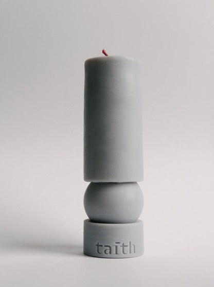 A Ninu Grey Candle by Taith sits atop a spherical base. The base, which is part of the same piece, has the word "taith" inscribed on it. Made from natural soy wax, this eco-conscious candle's minimalist design is emphasized by the plain, light-colored background.