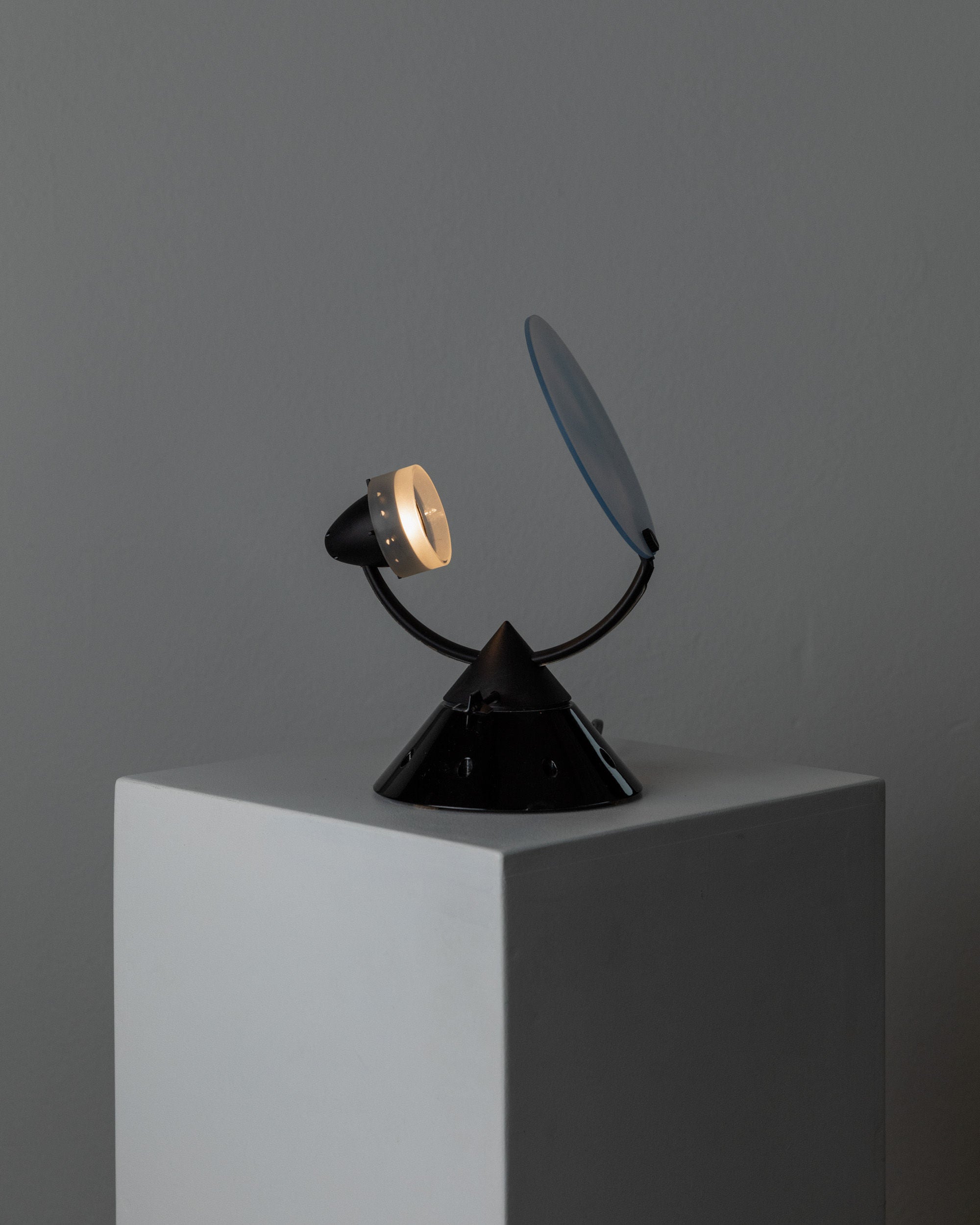 The Table Lamp by Lamperti, model Sestante (Italy, 1980s) from Bottega Jacobs features a geometric black form with a circular light and blue-tinted disc, displayed on a white pedestal against a gray wall—a sleek, minimalist Italian design.