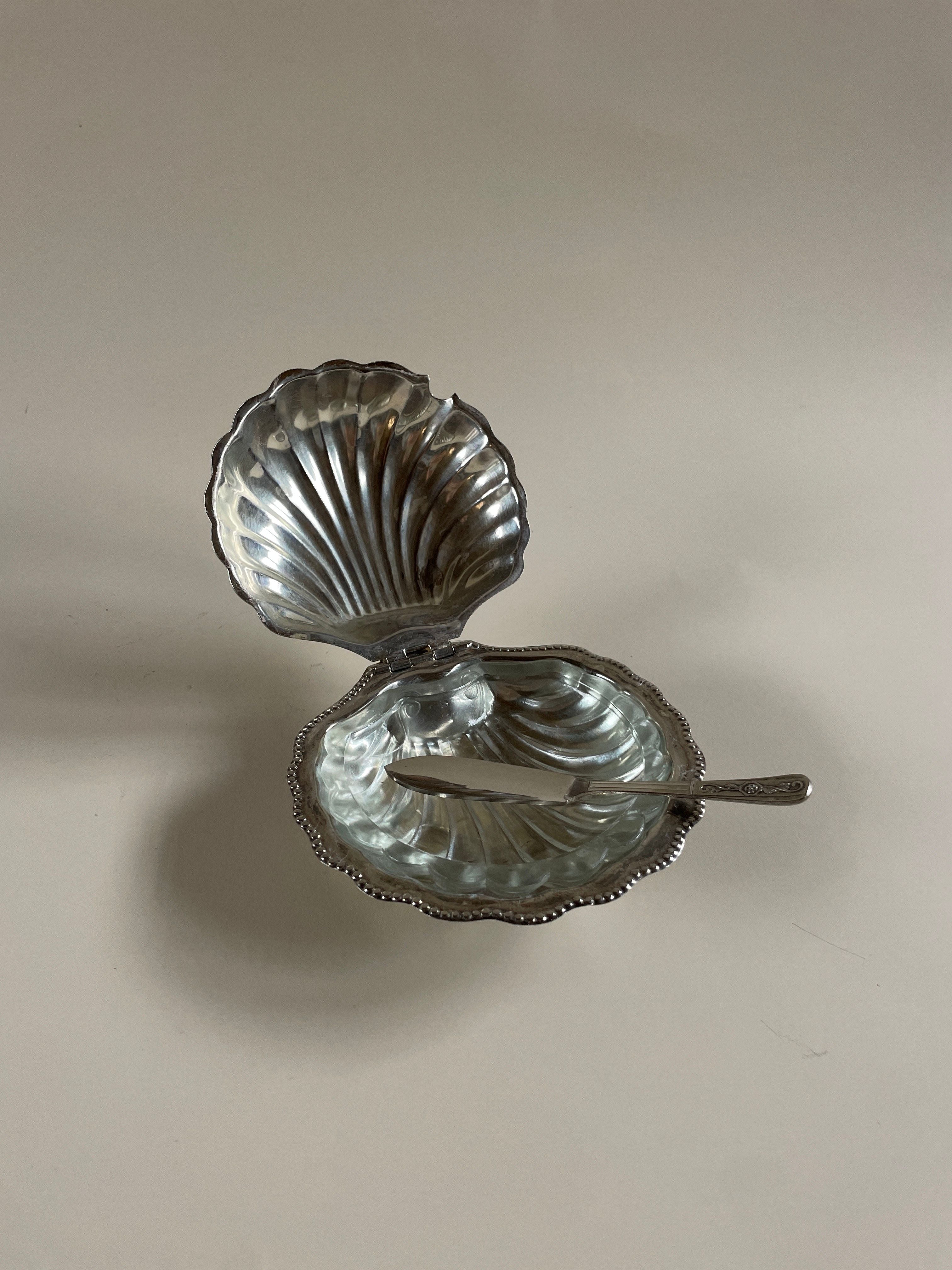 A Vintage Plated Shell Butter Dish by Maud Vaughan, complete with a small spoon and vintage butter knife, rests on a neutral background. Its intricate design resembles a scallop shell with detailed ridges and a glossy silver-plated finish.