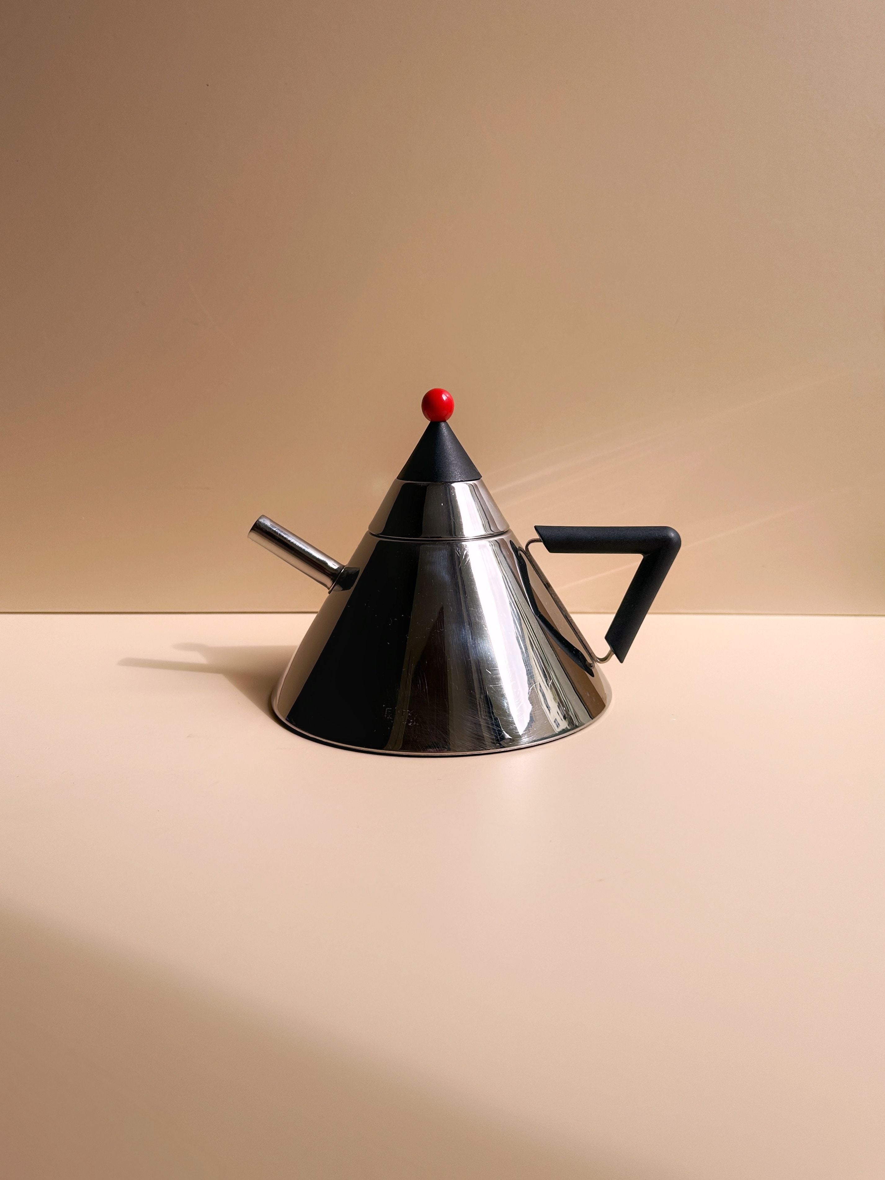 A modern, metallic Japanese Postmodern Steel Tea Pot from Dodo Vintage sits on a flat beige surface. Featuring a conical design with a small spout on one side and a black triangular handle on the other, it epitomizes postmodern design. The lid is topped with a small red sphere, blending seamlessly into the beige background.