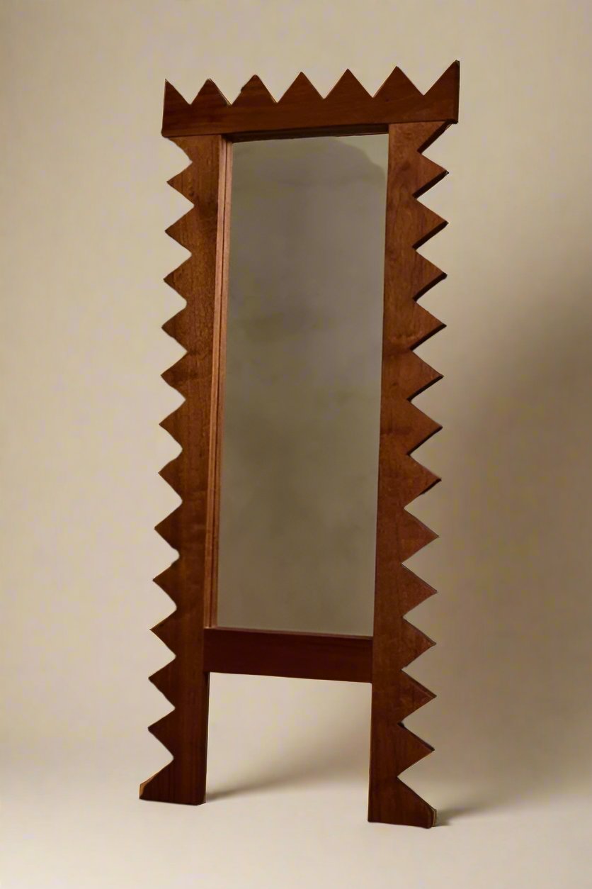 The Jura Mirror from Ah Um Design Studio, with its tall, rectangular form and solid mahogany frame featuring a jagged, zigzag edge, stands against a backdrop of beige curtains. Flanked by a pair of candlesticks and a potted plant on a wooden table, it introduces rustic, geometric forms that grant an artistic touch to the room.