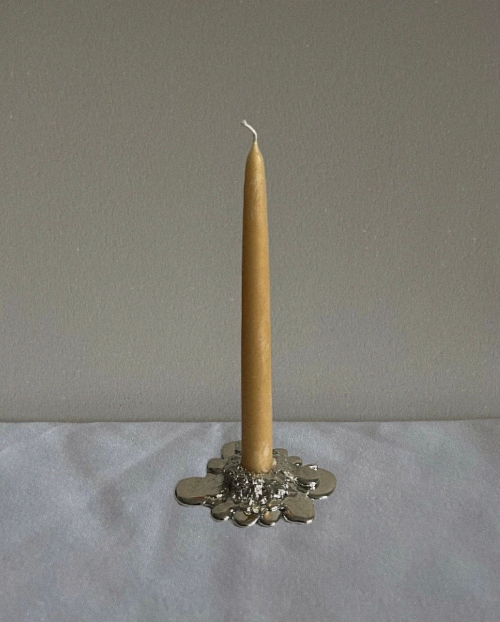A single, unlit beige candle stands upright in Studio Formbart's The Tin Collection / Melt Candle Holder, hand cast by Swedish artisans, set on a white surface against a plain gray wall.