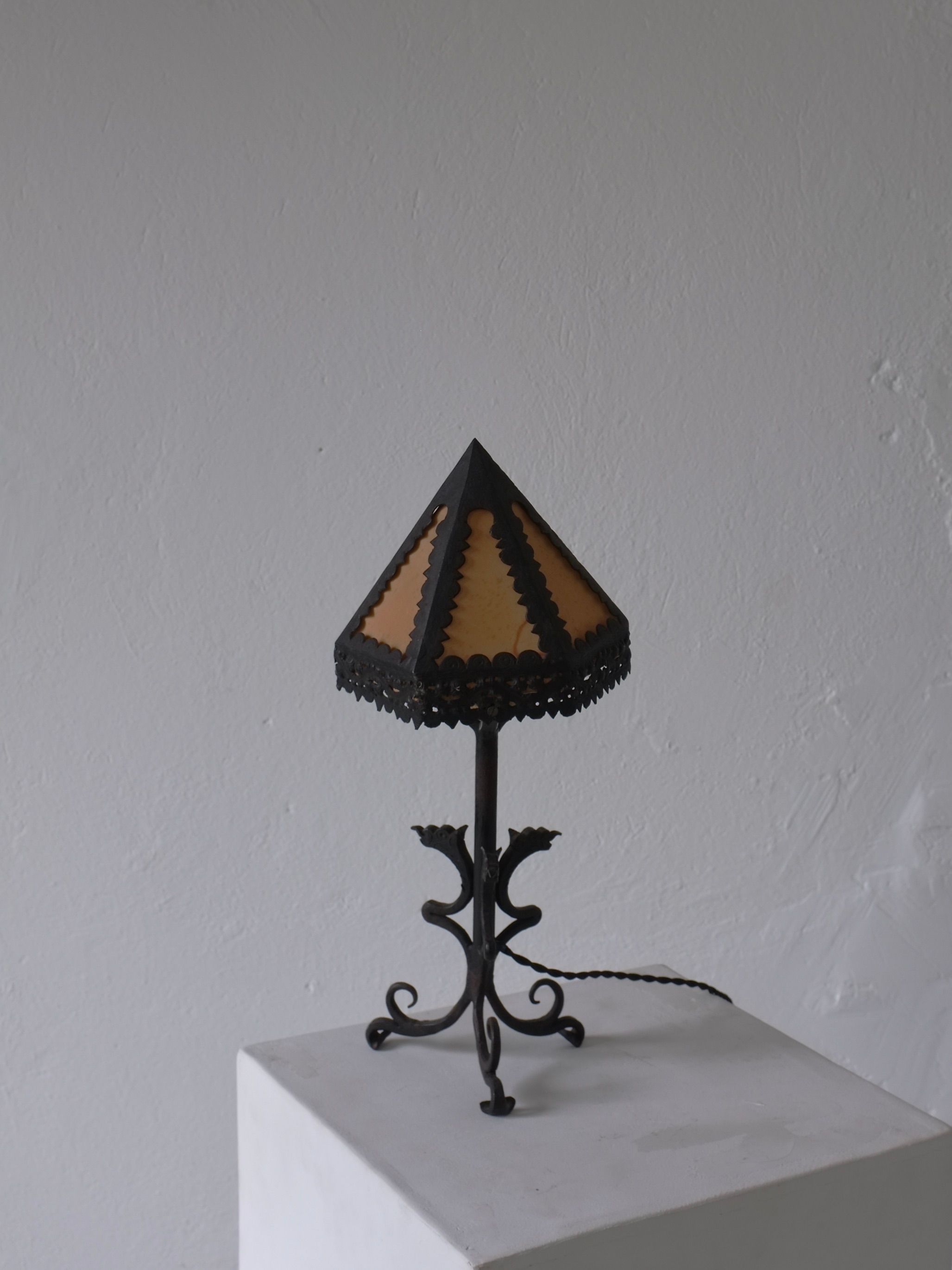 The Veter Vintage Wrought Iron Dragon Table Lamp, Italy 1910s, features a brown and orange stained glass shade and an ornate black metal base, displayed on a white pedestal against a light gray wall.