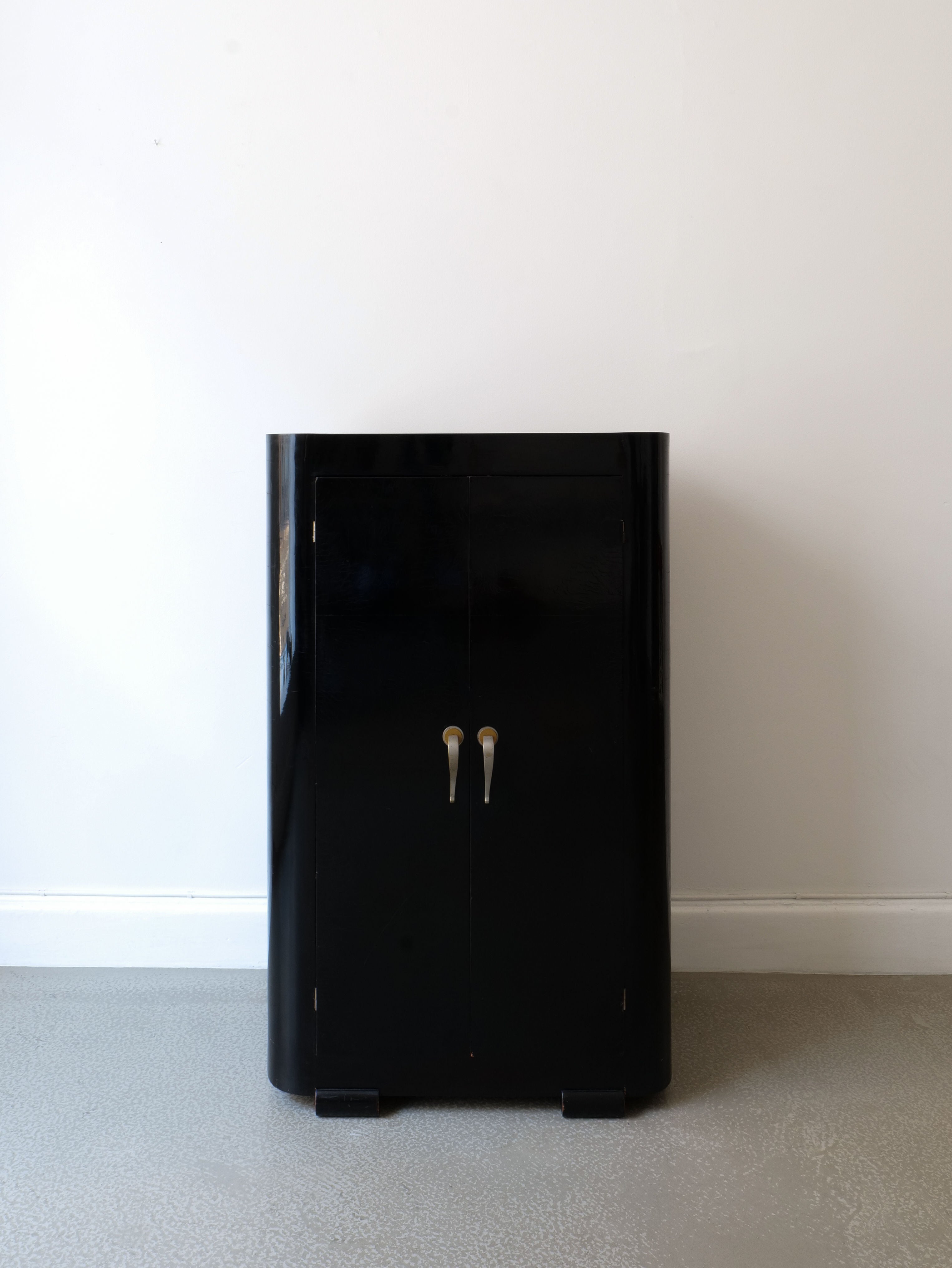 The French Deco Cabinet 1930s by Collection apart stands against a plain white wall. Featuring a sleek Art Deco design with a black lacquer finish, it has two vertical doors with metallic handles and rests on a speckled gray floor, reminiscent of French 1930s style.