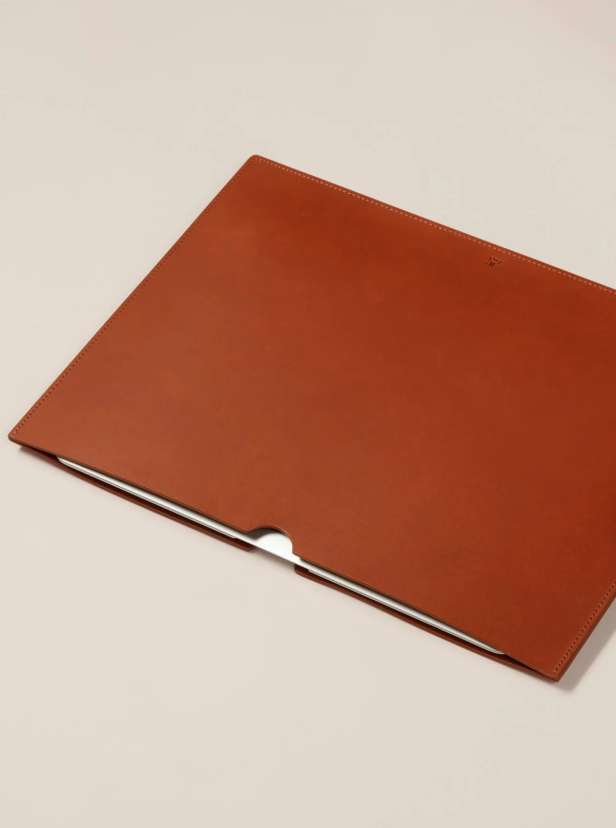 A luxury Paradise Row Leather Laptop Sleeve is placed on a light beige surface. Crafted from vegetable tanned leather, it boasts a minimalist design with a convenient notch for easy access. The smooth texture and visible stitching highlight the elegance of its ethically sourced materials.