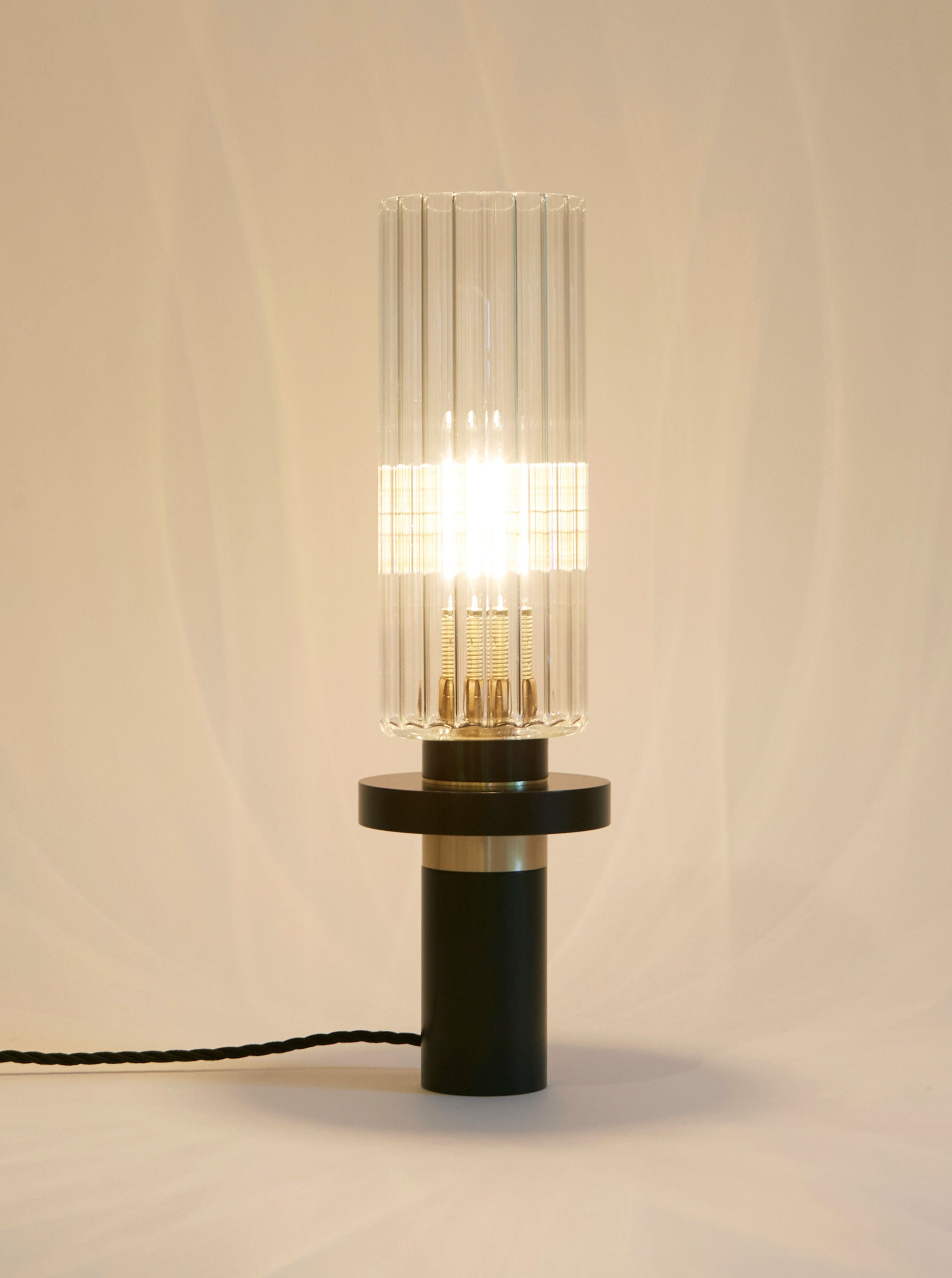 A modern Alexandre table lamp by Marine Breynaert with a striped transparent glass lampshade and a black base, illuminated and casting a warm glow against a neutral background.
