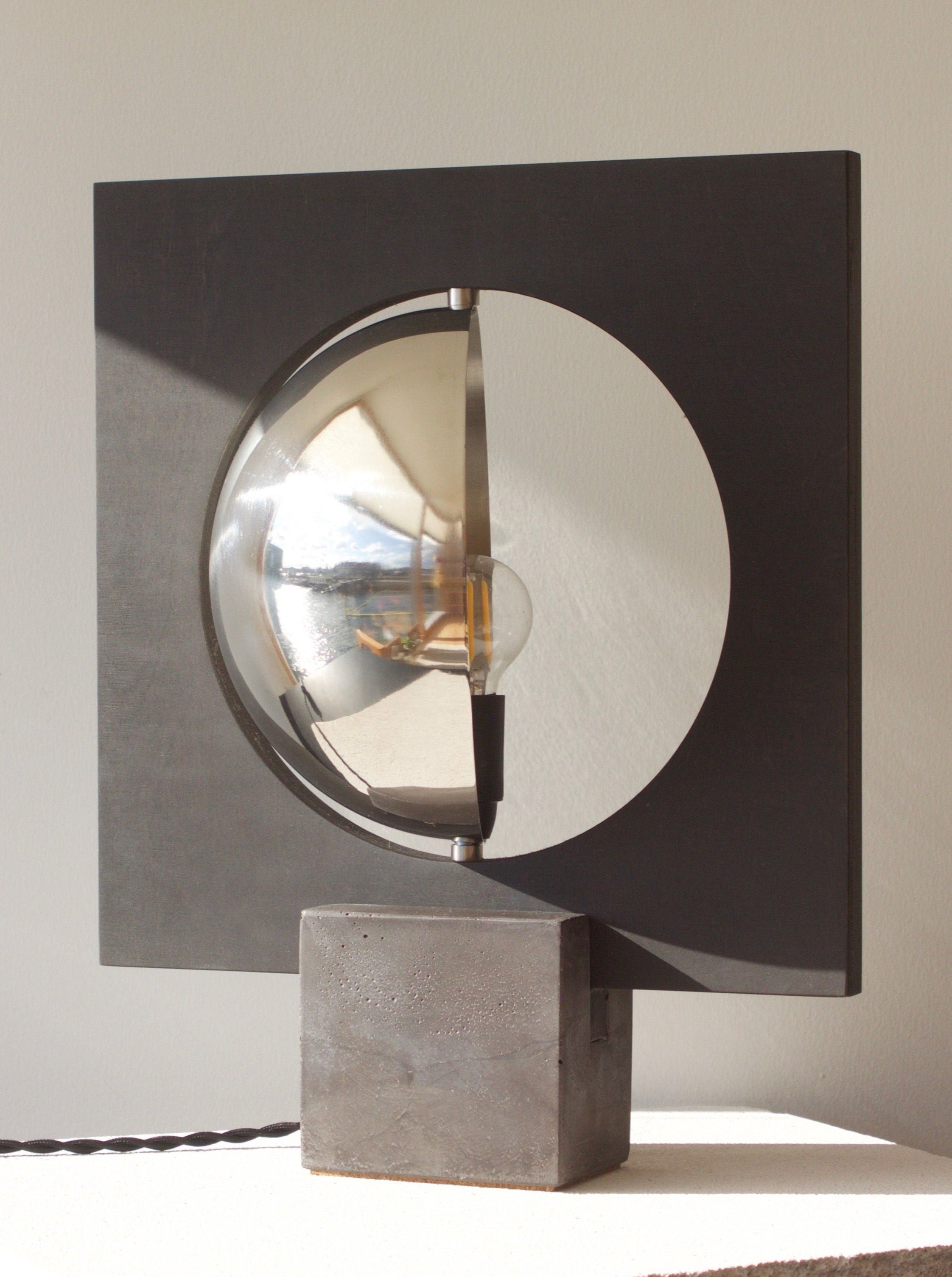 A modern floor lamp featuring a circular mirror centered in a square, black frame, mounted on a gray concrete base, placed on a white surface against a neutral wall, the Galileo 2.0 by Edoardo Lietti Studio.