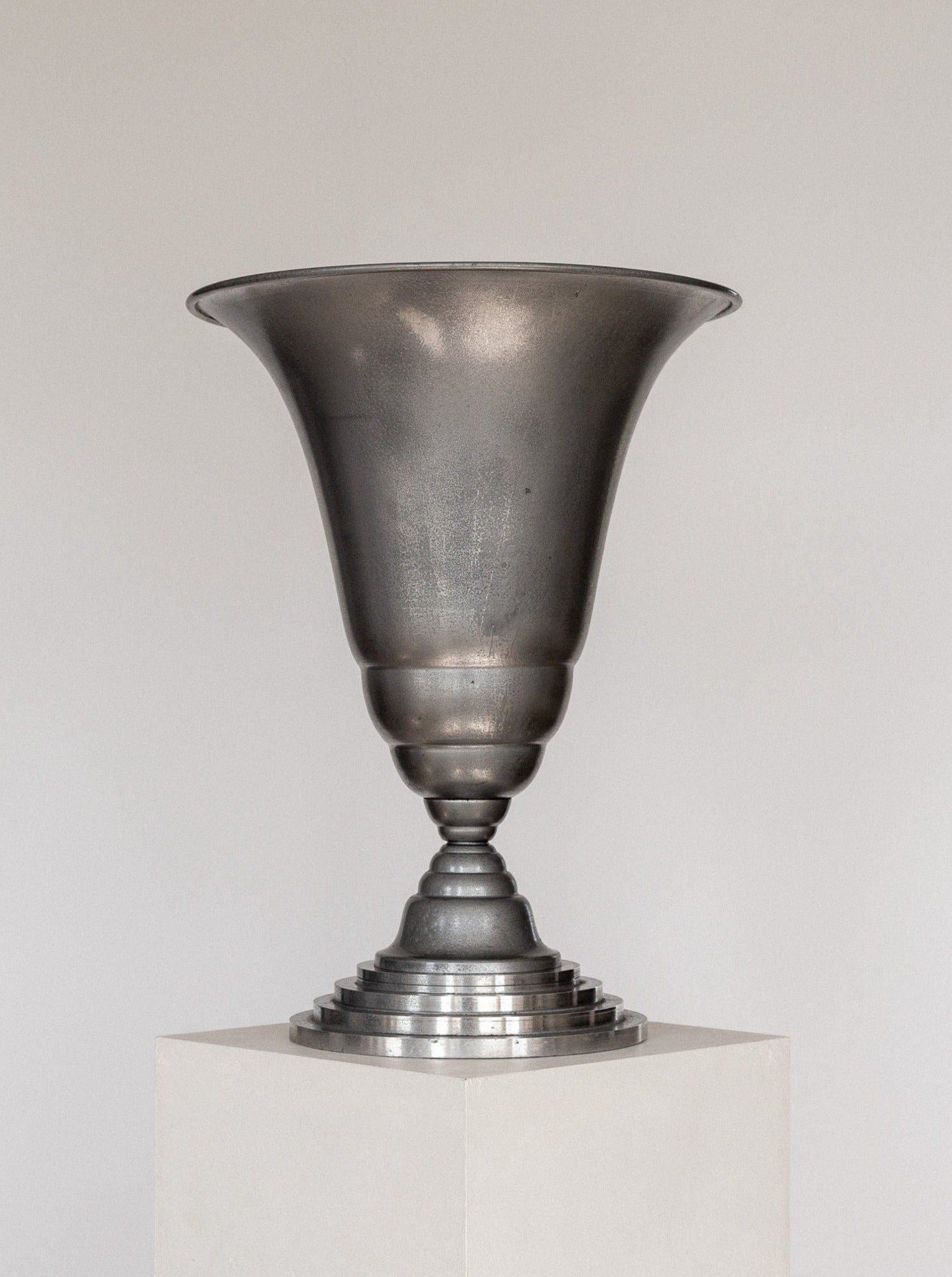 The Art Deco Torchière Table Lamp by Spigel, featuring a wide, flared brim, a narrow base, and a tiered stand, sits atop a white pedestal against a plain background. The French-designed lamp has a slightly tarnished, brushed finish that exudes vintage elegance.