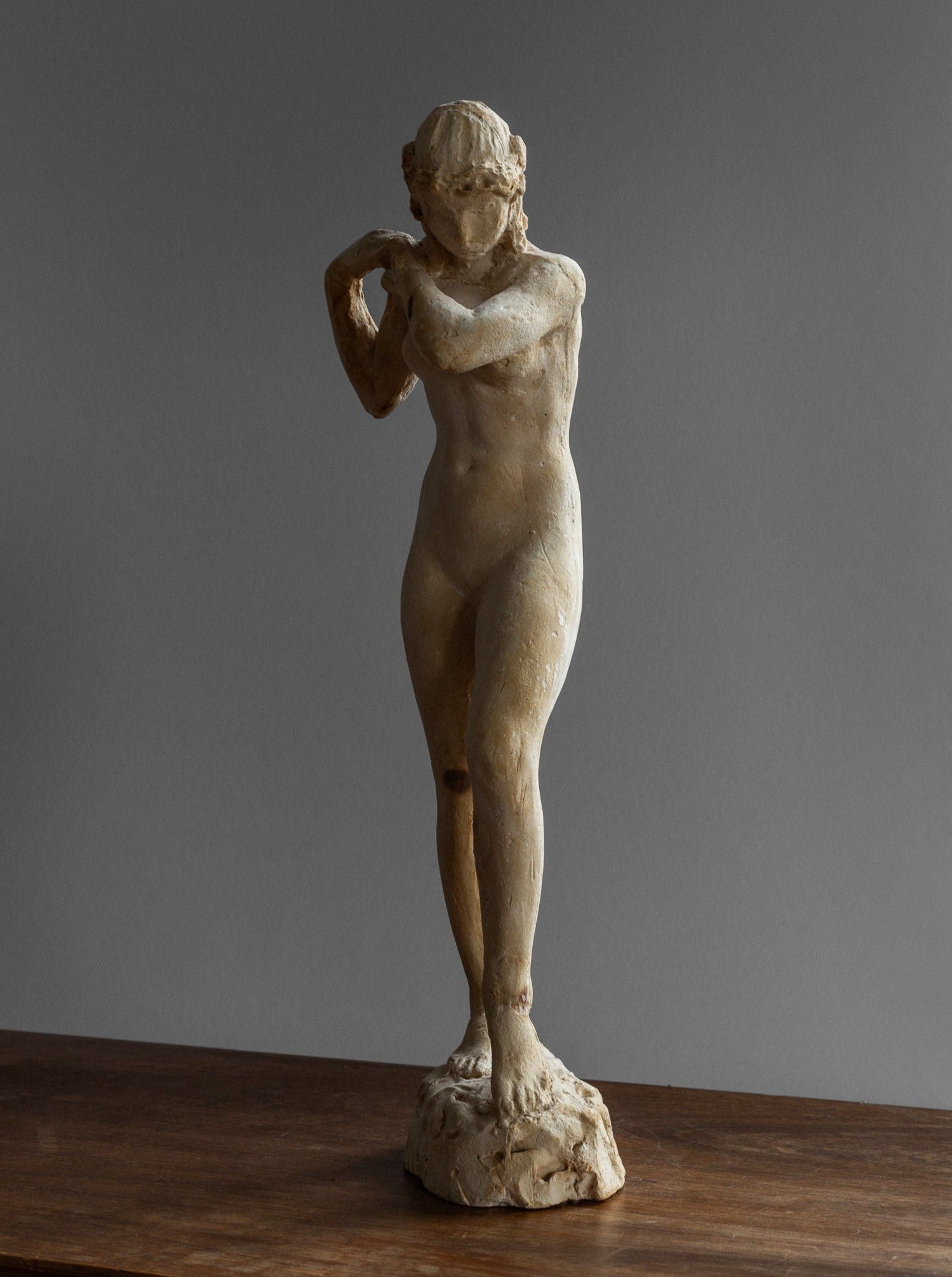 The Vintage Neoclassical Sculpture by Spigel, originating from Sweden and crafted in the 1920s-30s, features a standing naked figure with a body slightly bent at the knees and arms raised near its neck. The delicate details of this plaster statue are presented on a wooden surface against a plain gray background.