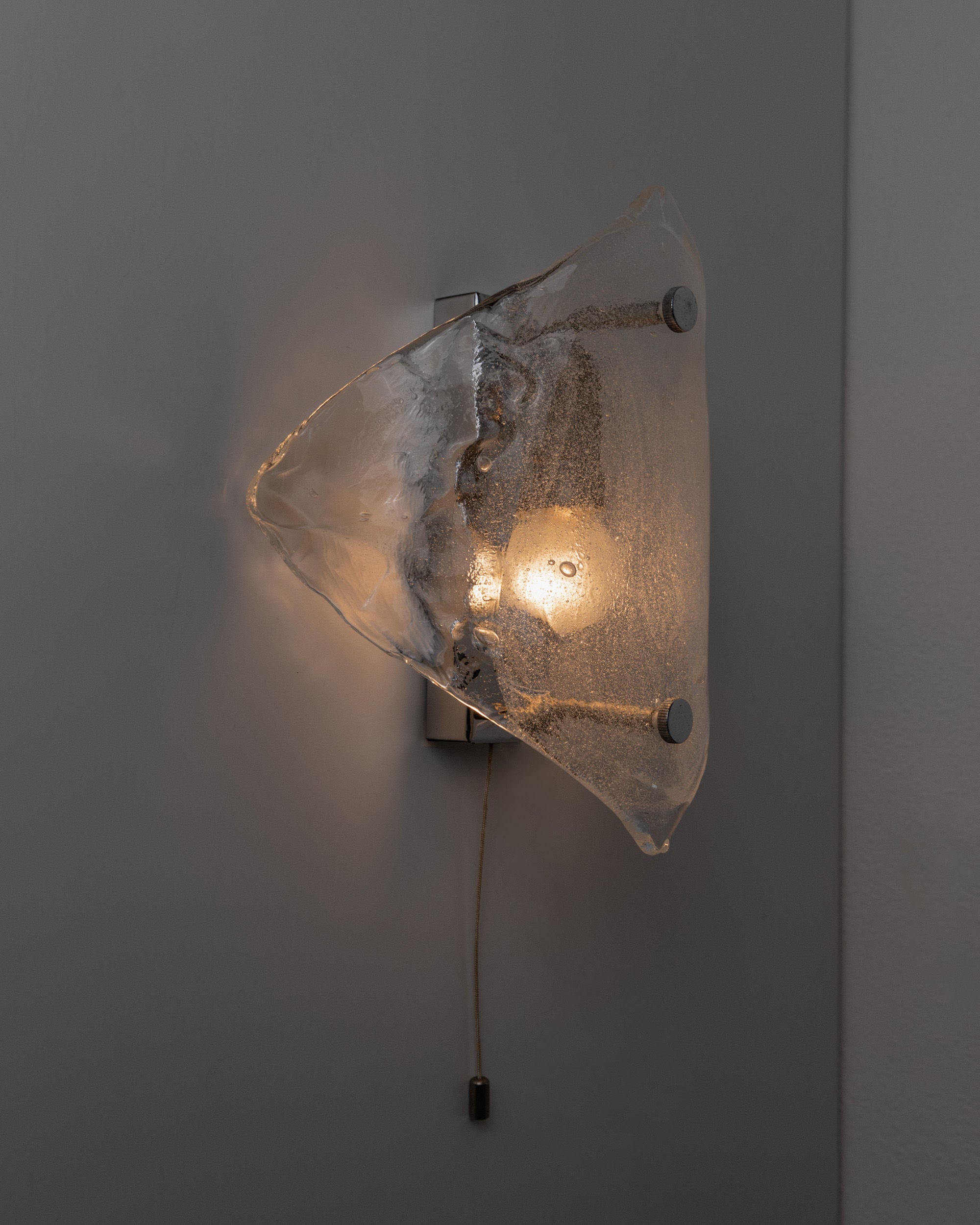 The Vintage Murano Glass Wall Lamp by Kalmar from Bottega Jacobs features a space-age design with an abstract, translucent glass shade. A warm glow emanates from the bulb held by two metal rods, and it's operated via a pull cord, embodying the elegance of handmade glass art.