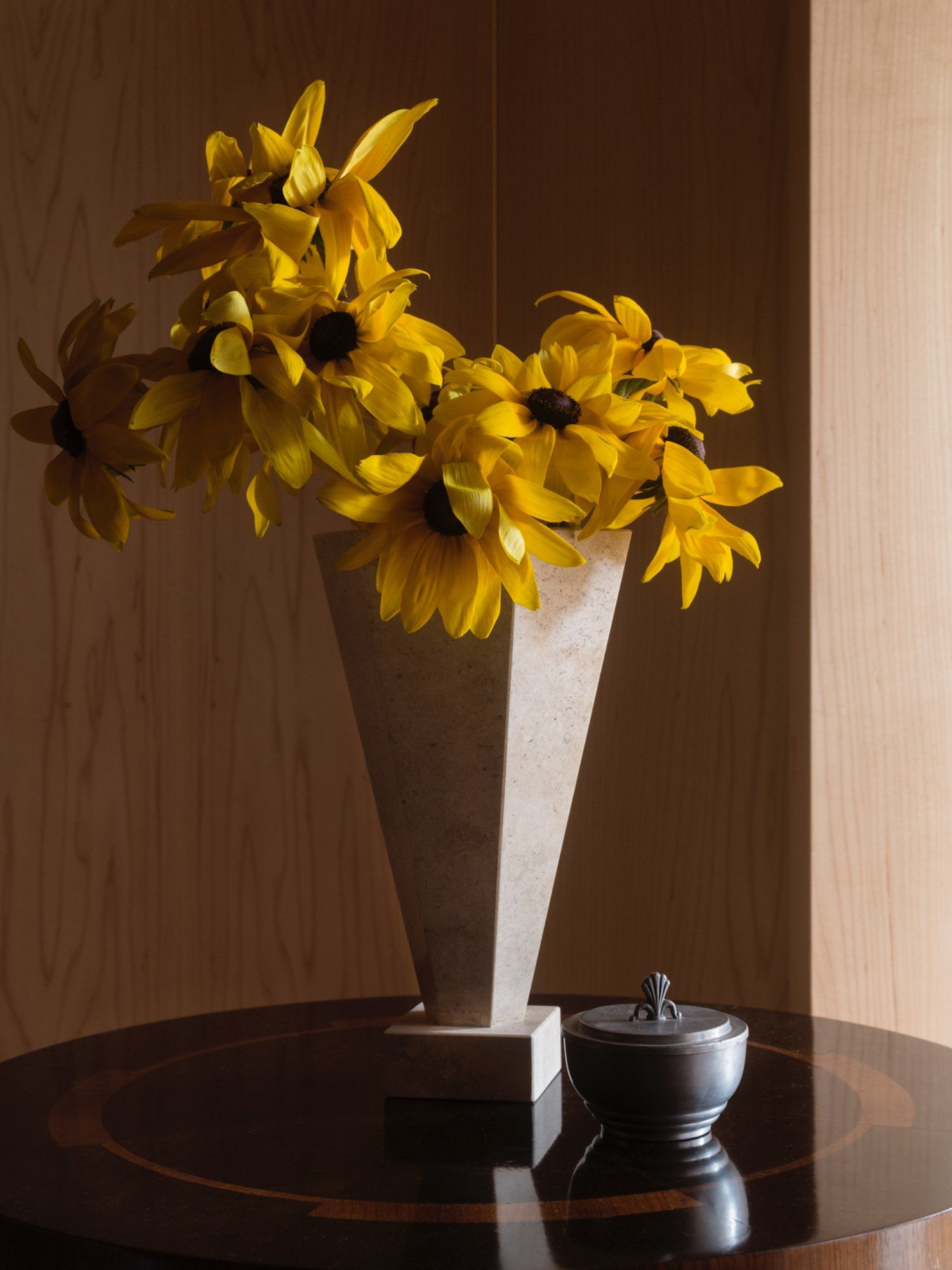 A Vase 01 Travertine from Studio Vraco, filled with vibrant yellow flowers, adorns a round wooden table. Beside the vase rests a small, dark metal container with a decorative handle from the TSDXSV Collection. The scene is set against light wood paneling that adds a warm ambiance.