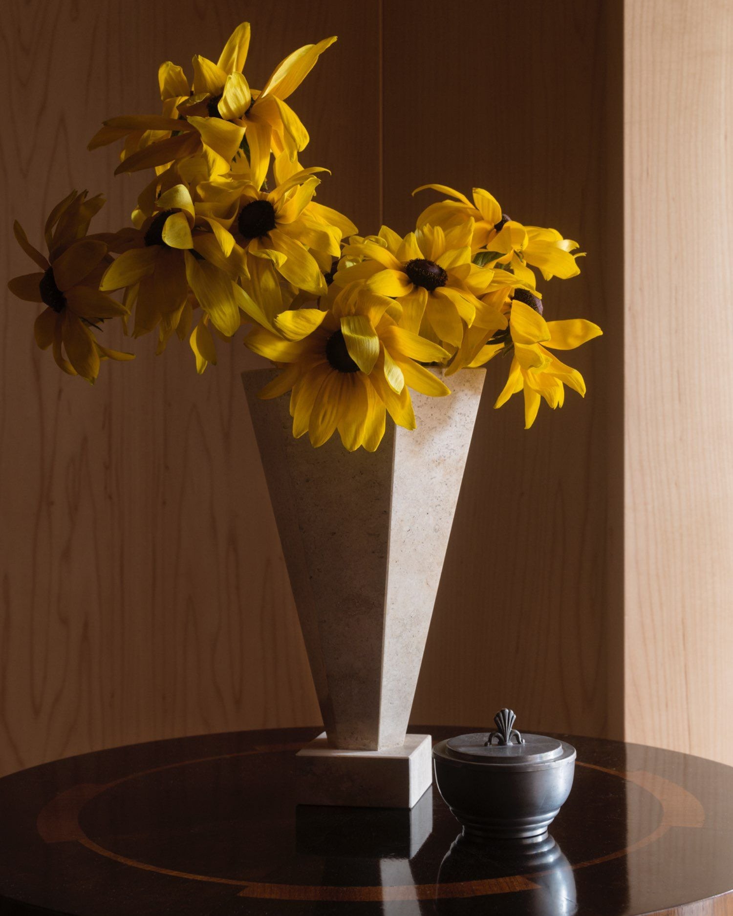 A Vase 01 Travertine from Studio Vraco, filled with vibrant yellow flowers, adorns a round wooden table. Beside the vase rests a small, dark metal container with a decorative handle from the TSDXSV Collection. The scene is set against light wood paneling that adds a warm ambiance.