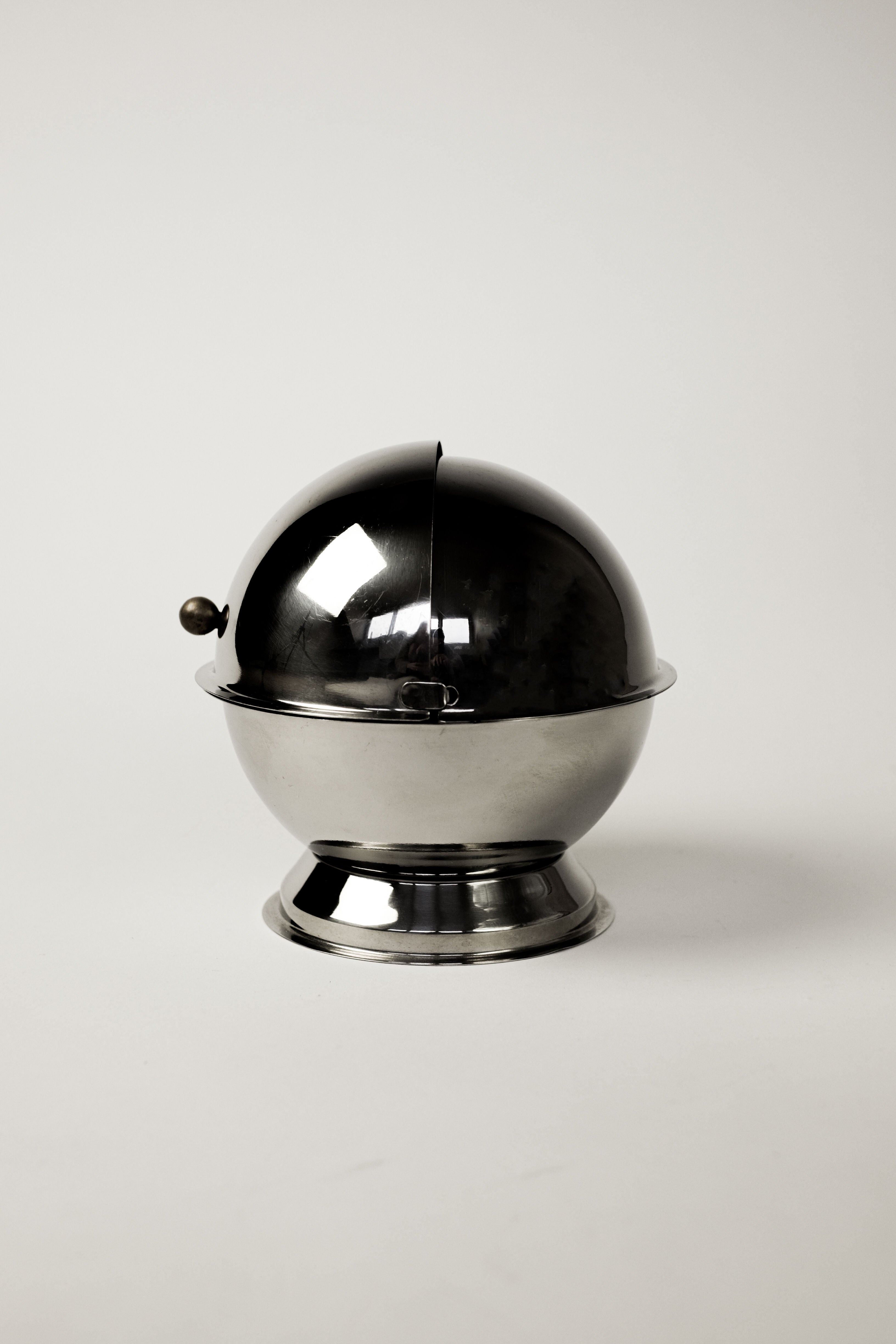 The Circle Sugar Bowl by septembre studios features a shiny, spherical design with a round lid and small handle. Sitting on a flat base against a plain background, its reflective surface and vintage appeal cast subtle shadows.