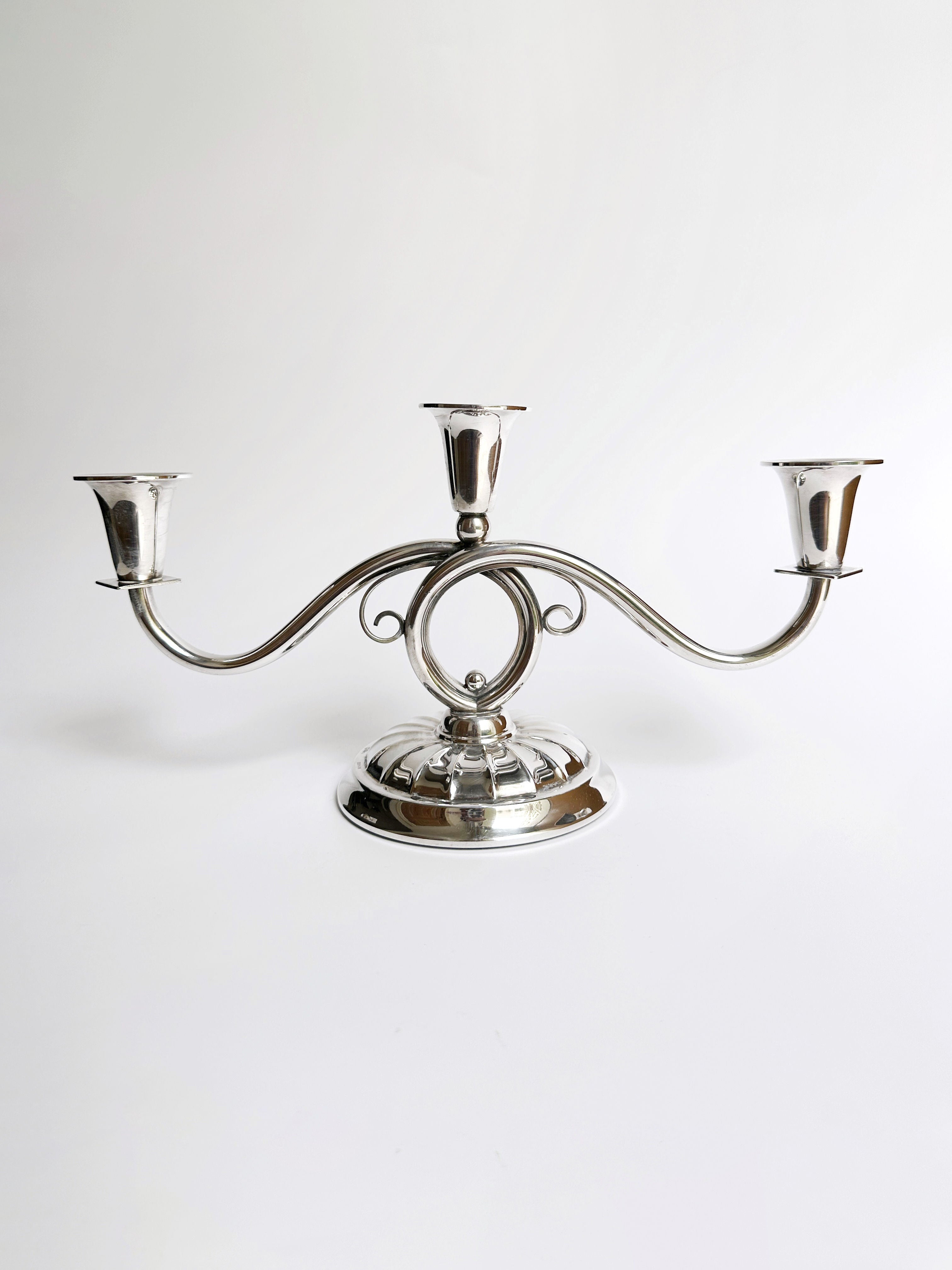A Danish Mid Century Candleholder by Dodo Vintage, crafted in silver with an elegant, symmetrical three-arm design. The center arm boasts a circular loop, and the rounded base is adorned with decorative filigree details. This exquisite piece is showcased against a plain white background.