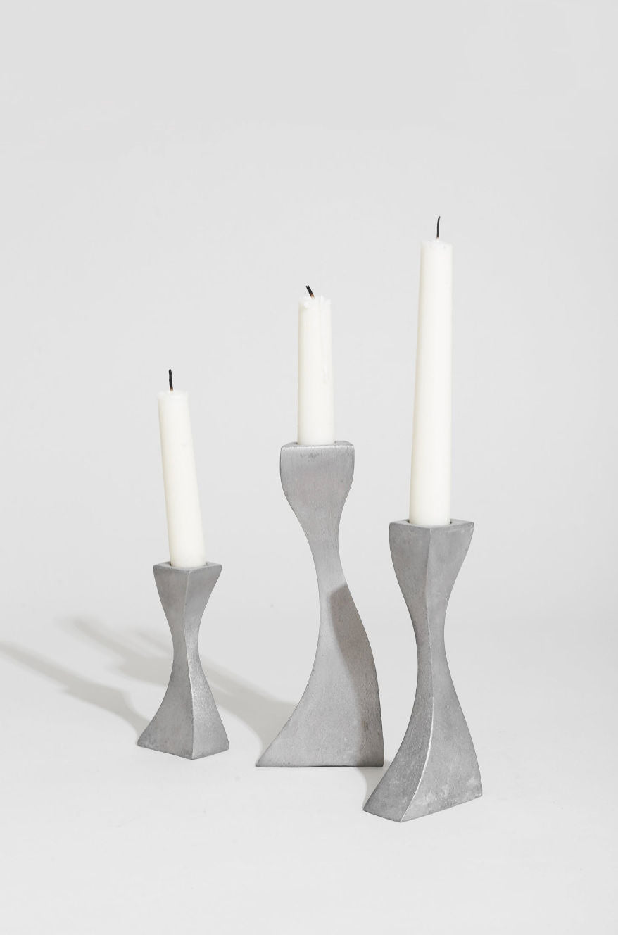 The Villa Wavy Candleholder Set features a sleek metal trio with hourglass shapes and tapered edges, displaying white candles with blackened wicks against a simple white background.