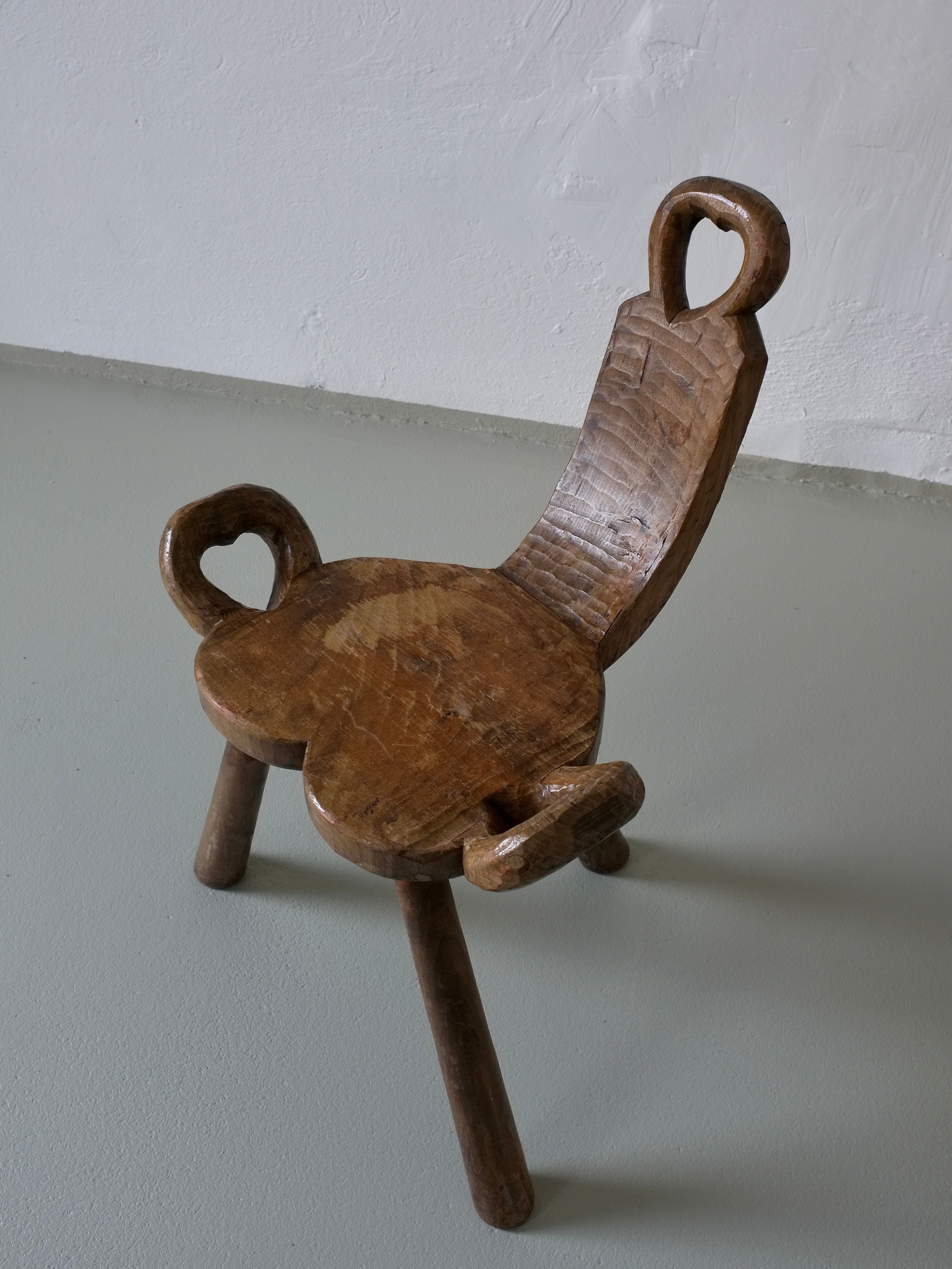 This Veter Vintage piece, the Vintage Brutalist Carved Tripod Stool, features a carved wooden design with a unique heart-shaped backrest and seat. Its three-legged structure emits handcrafted charm with a rustic finish, echoing 1960s Belgian furniture against a light-toned backdrop.