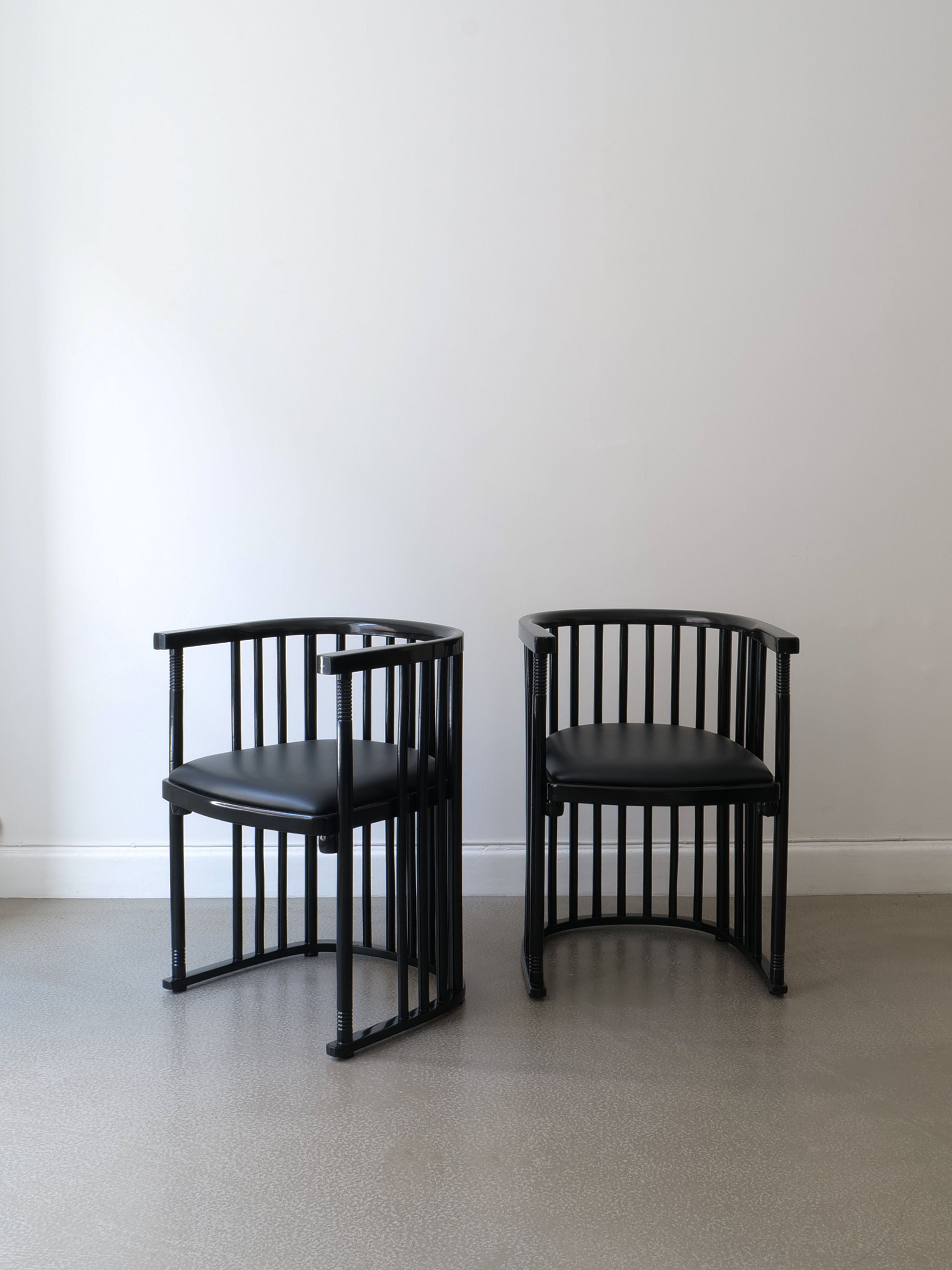 Two sleek and modern Bentwood Armchairs by Collection Apart, featuring Josef Hoffman's signature round backs and vertical slats, are positioned side by side on a smooth, light gray floor. The elegant contrast of their lacquered bentwood against the plain white wall adds a touch of sophistication to the scene.